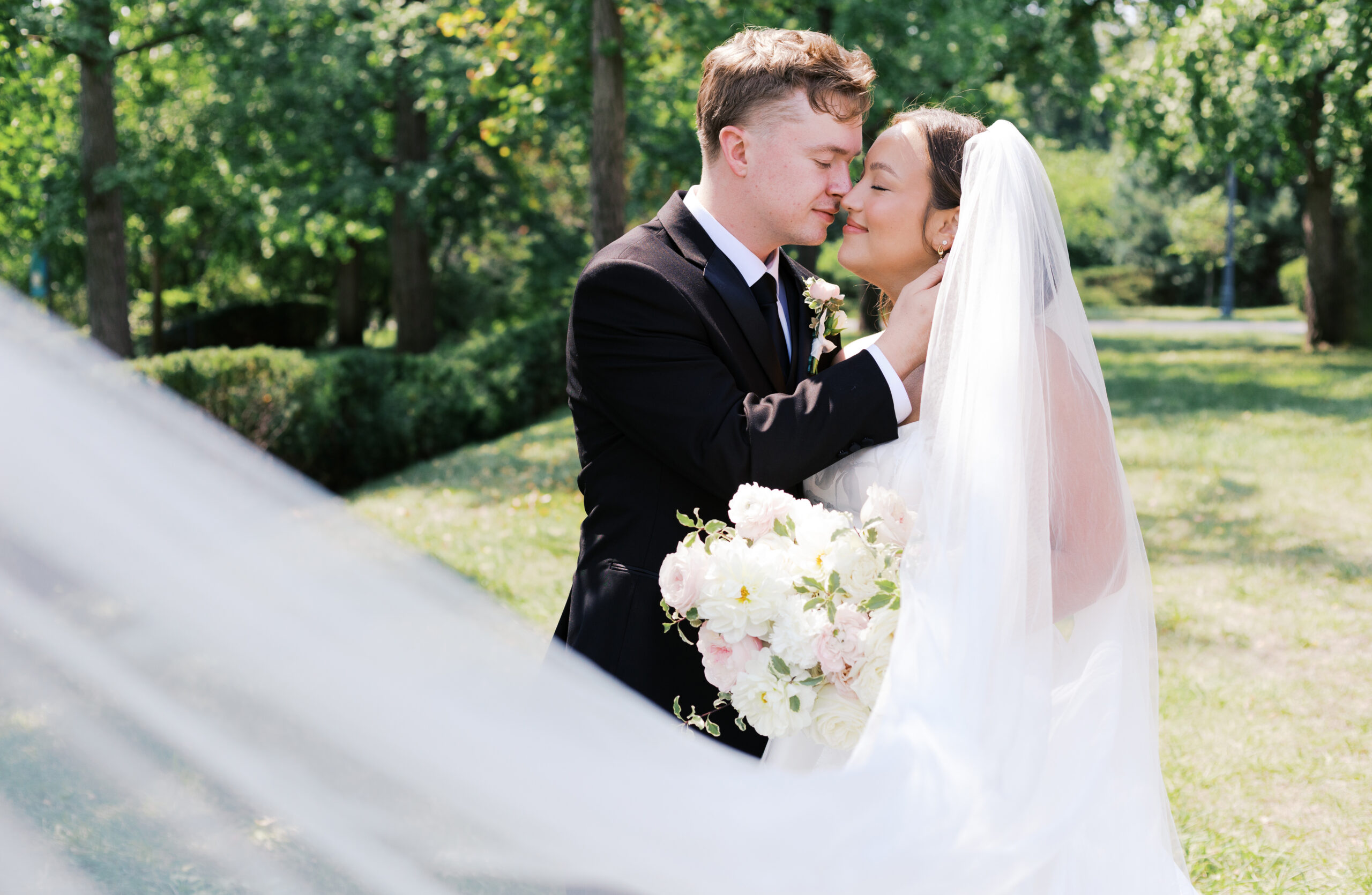 Bride and groom standing close in a sunlit park, holding a bouquet as the groom gently touches the bride’s neck.