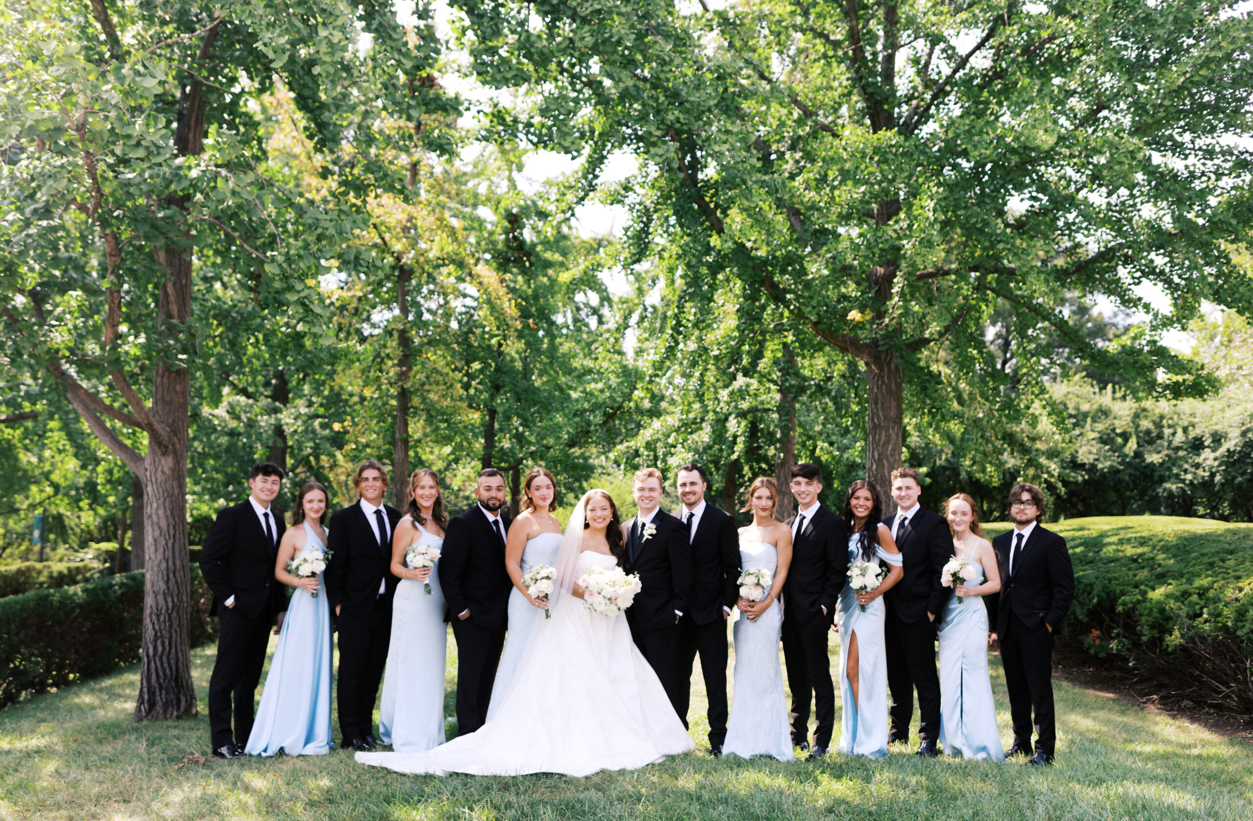 Wedding party standing in a line outdoors with the bride and groom centered beneath leafy trees.