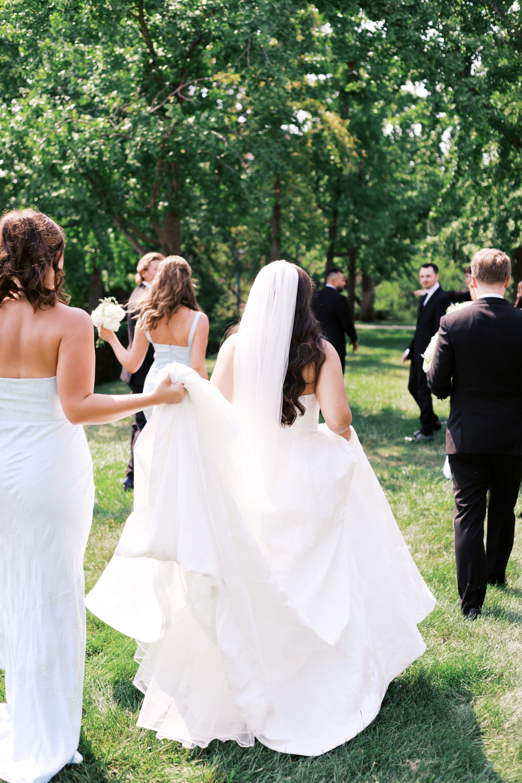 Bride walking away from the camera with her veil flowing, surrounded by the wedding party in a green park.