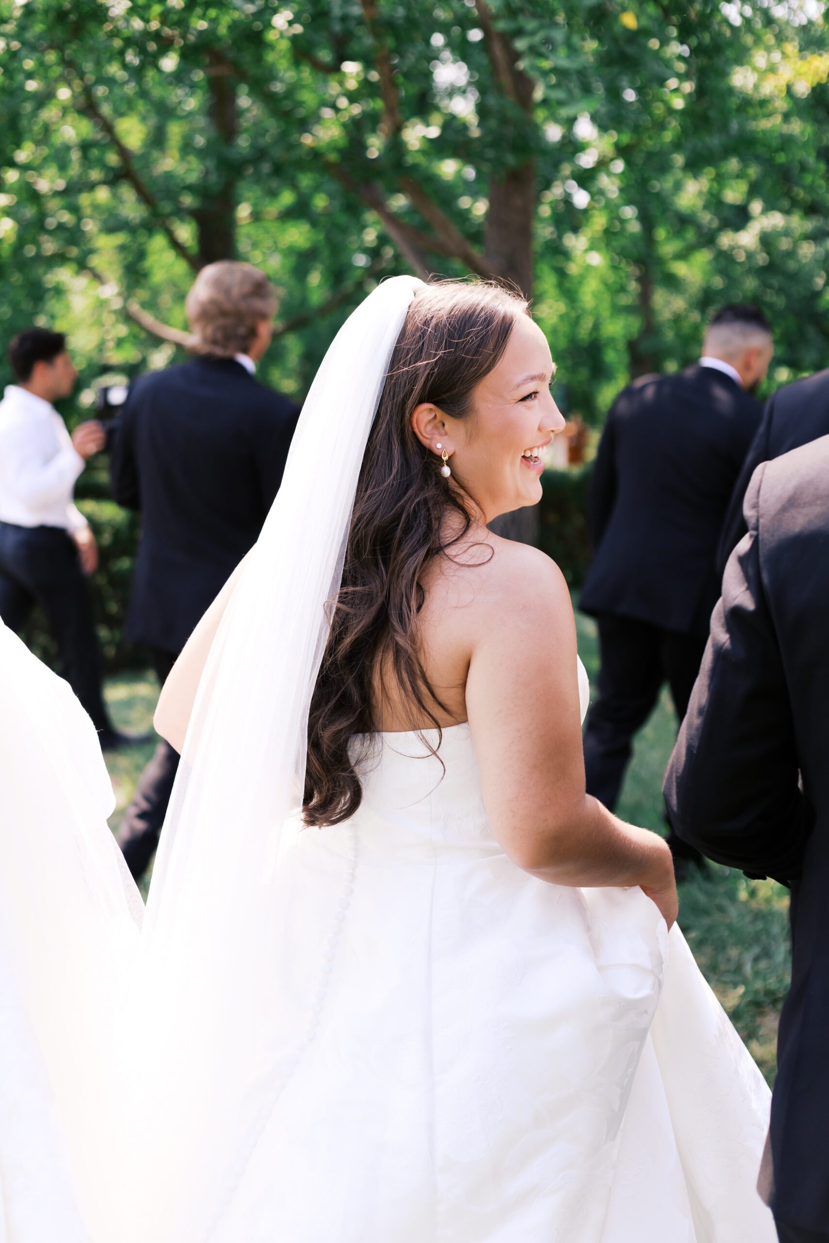 Candid side view of the bride smiling while walking outdoors, with wedding guests blurred in the background.