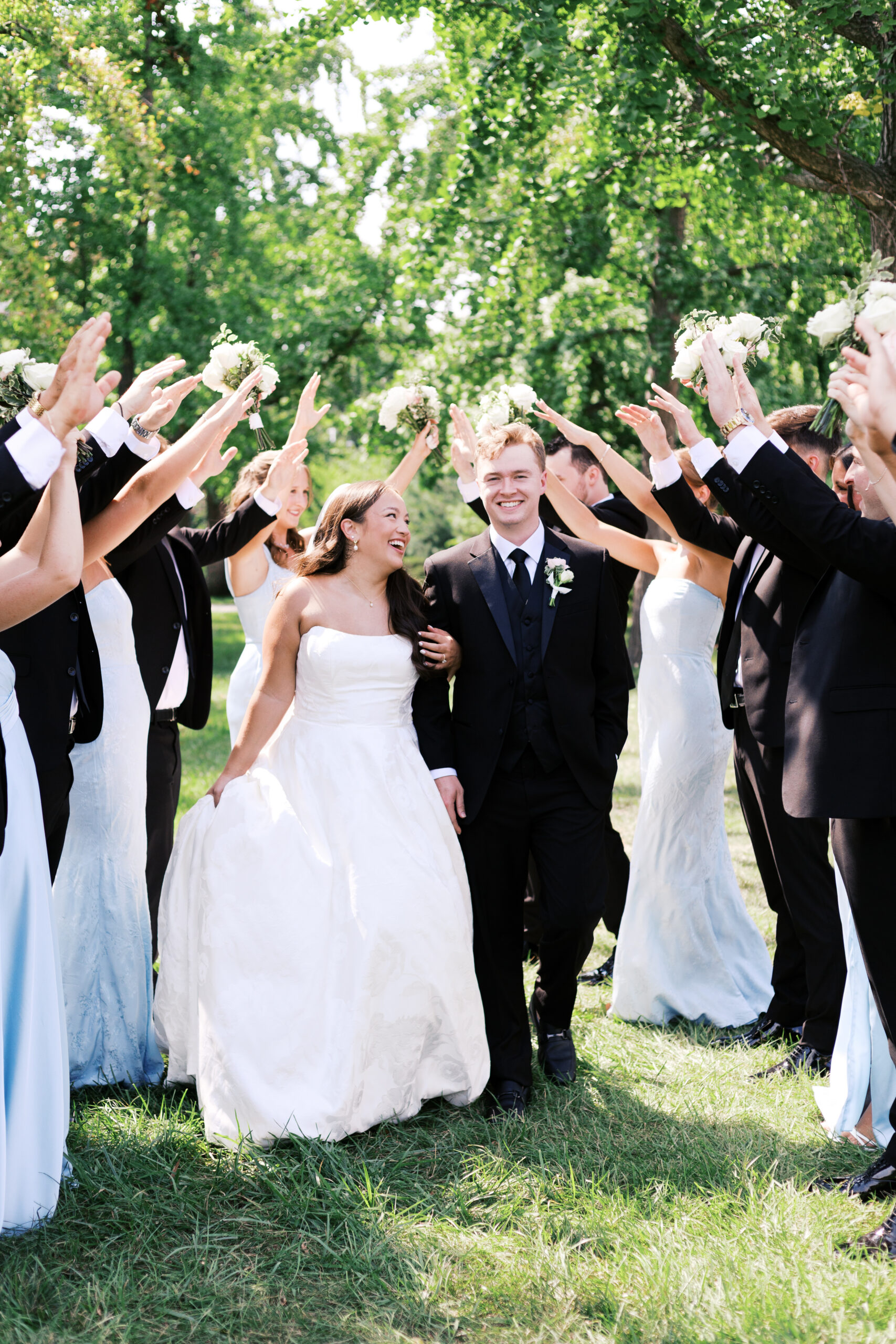 Bride and groom walking between raised arms of their wedding party holding bouquets in a celebratory outdoor moment.