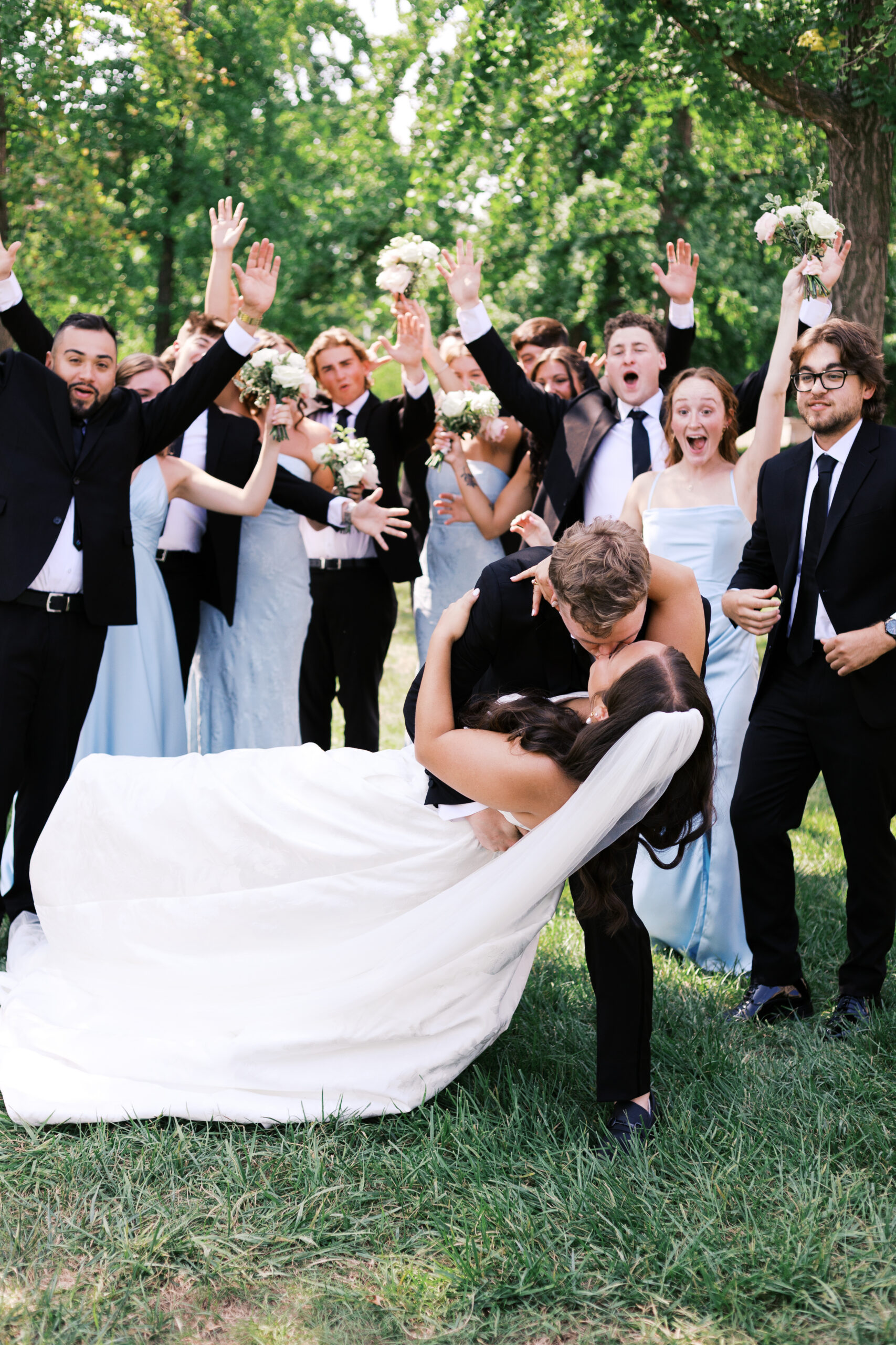 Bride dipped backward by the groom during a kiss as the wedding party cheers behind them in a green park.