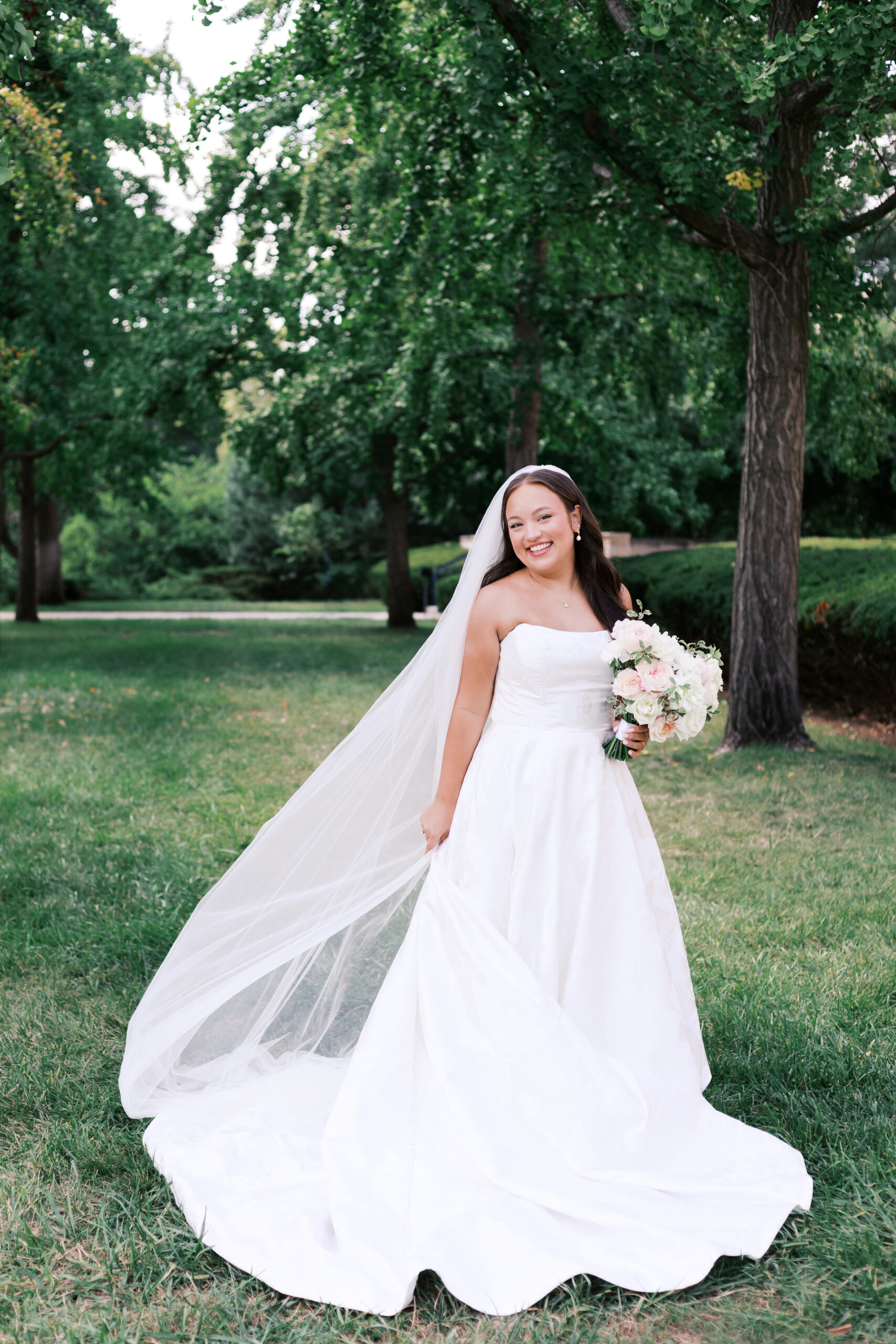 Bride standing alone in a grassy park, holding a bouquet with her long veil flowing behind her.