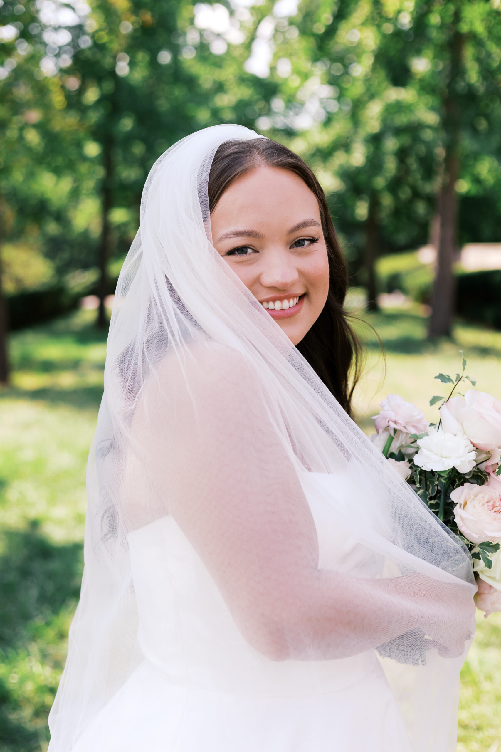Close-up portrait of the bride smiling over her shoulder, with her veil softly draped and greenery blurred behind her.