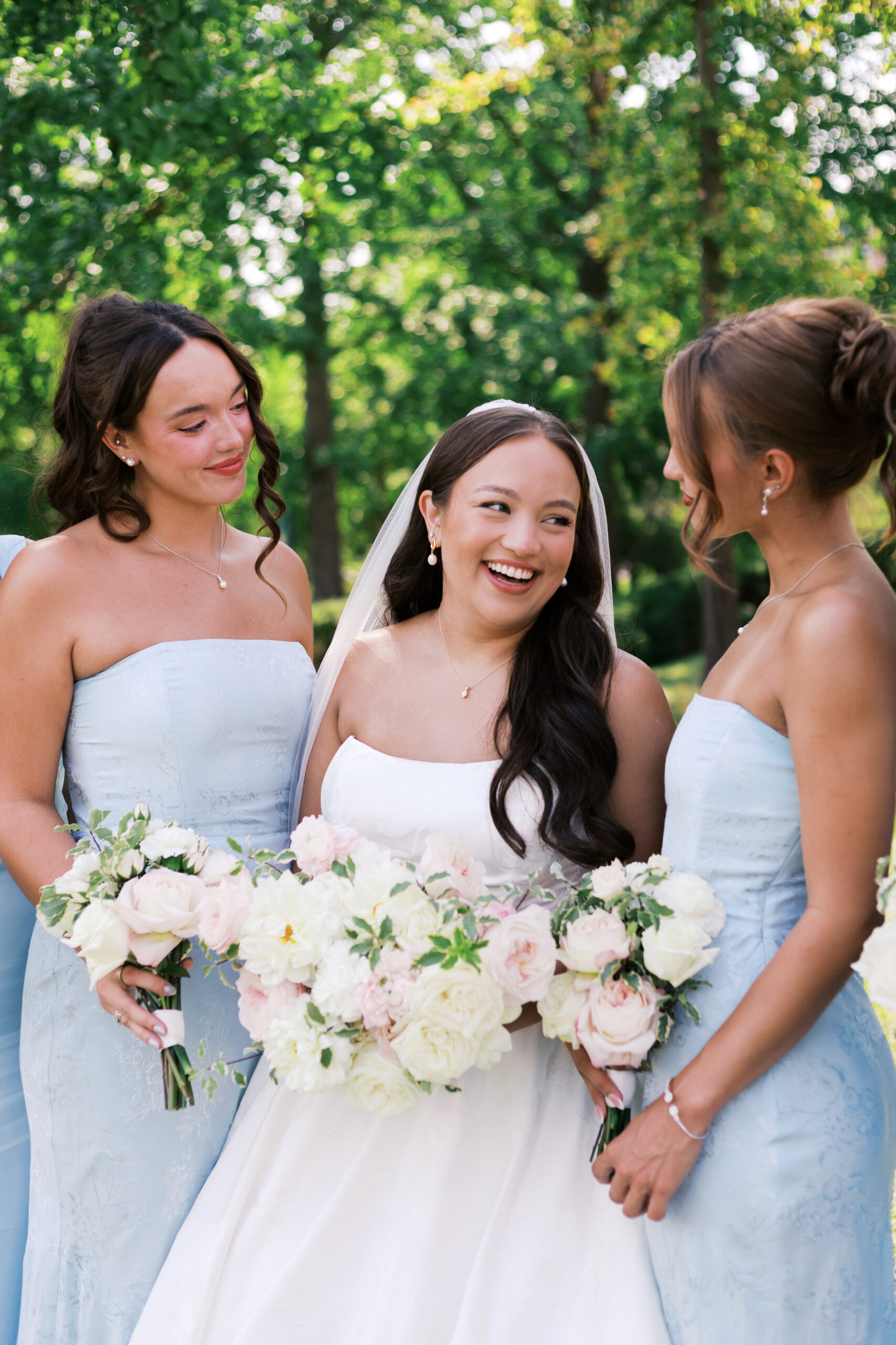 Bride laughing with two bridesmaids while holding a white and blush bouquet during outdoor wedding portraits.