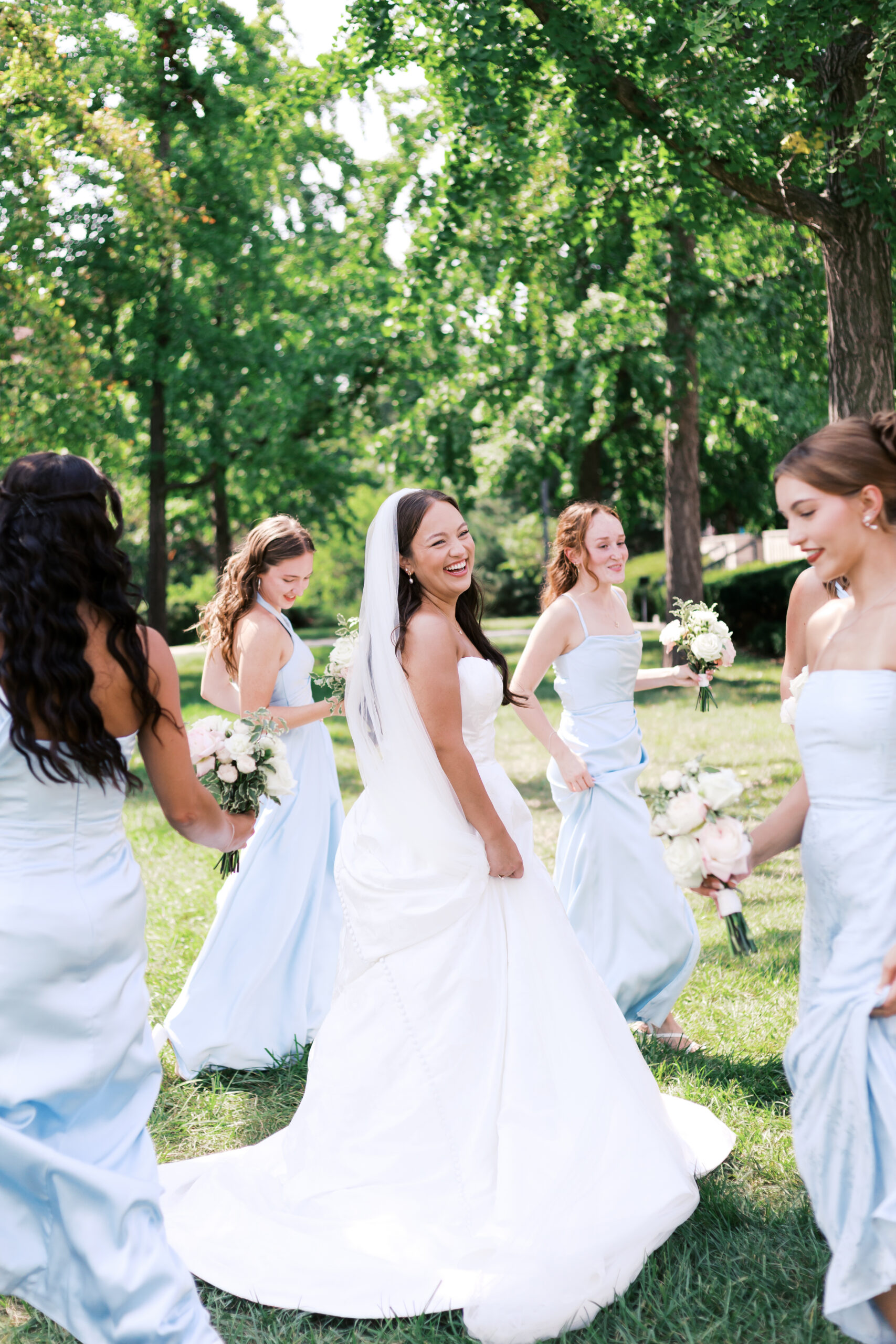 Smiling bride in white dress and veil walking with bridesmaids holding bouquets in a sunny park.