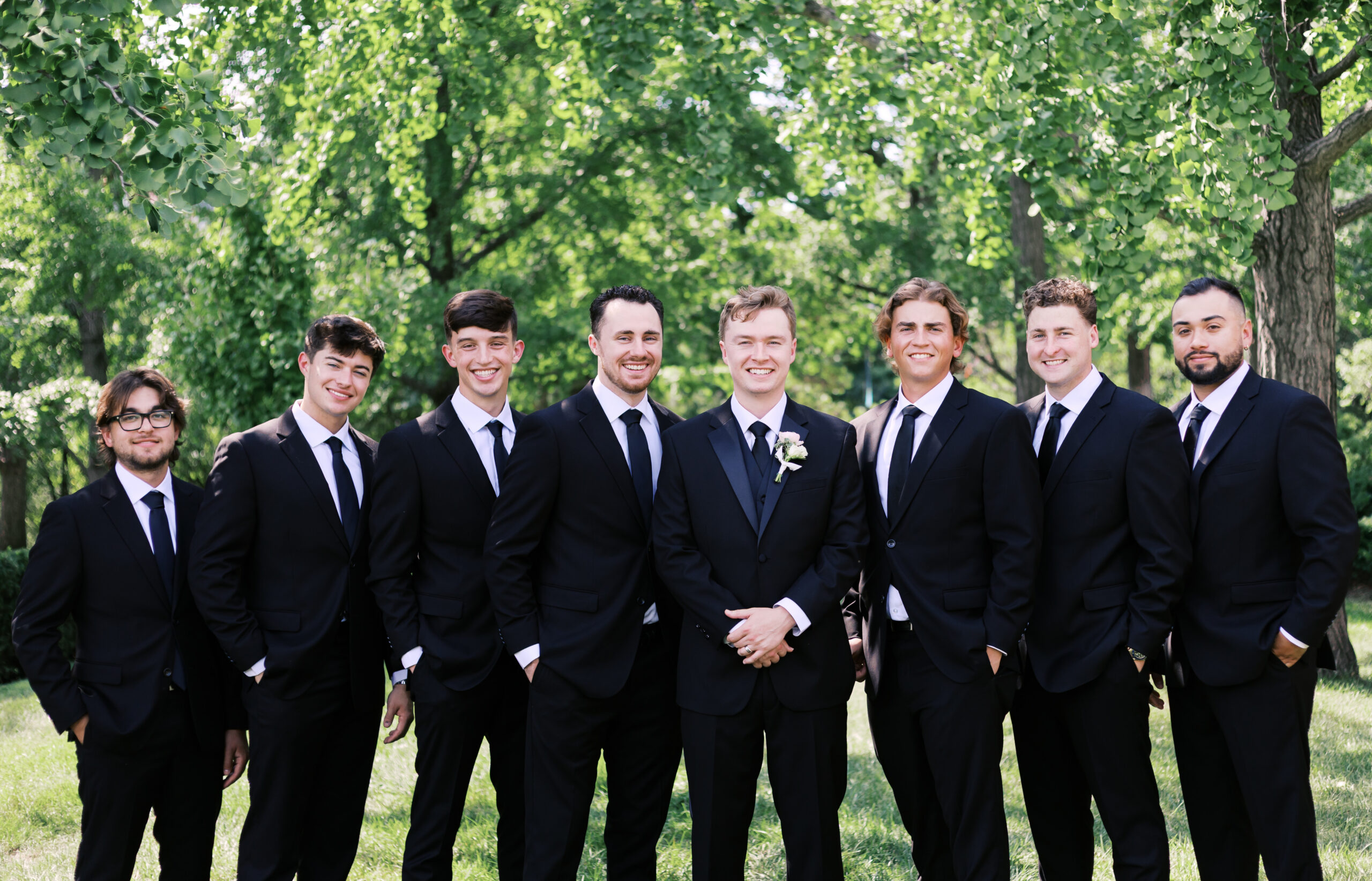Eight groomsmen pose in a line wearing matching black suits with white shirts and black ties, standing on grass with lush green trees behind them. The groom in the center-right wears a white floral boutonniere. The group smiles at the camera in a formal wedding party portrait.