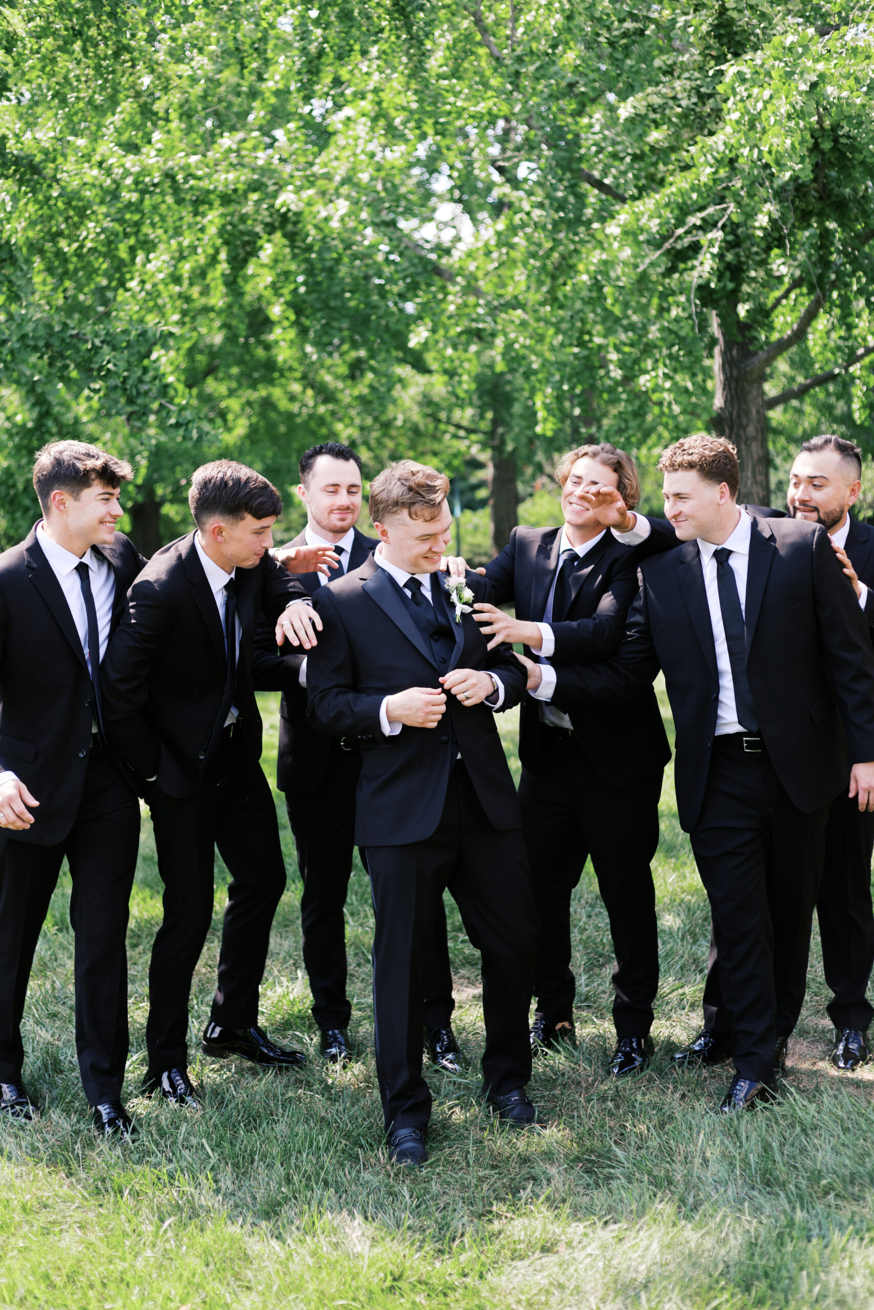Groom surrounded by groomsmen in black suits laughing and celebrating together outdoors.