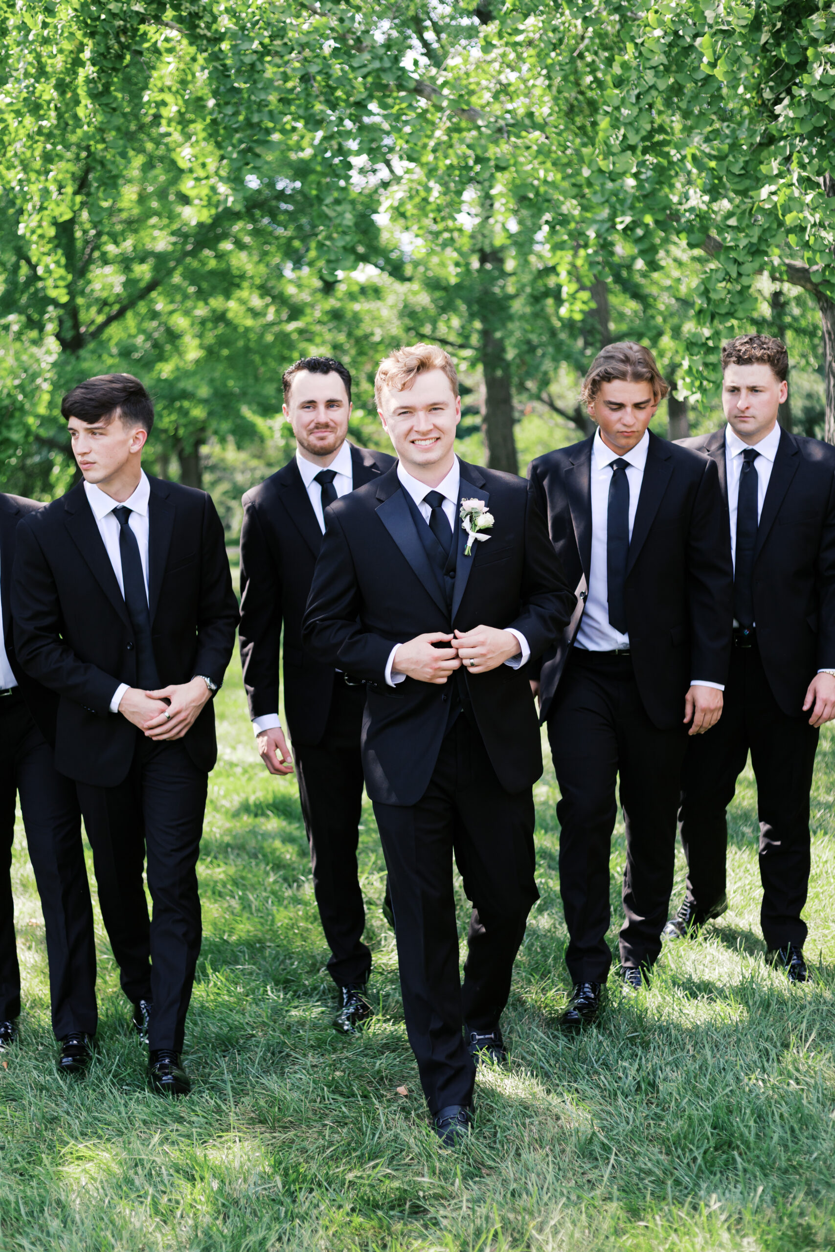 Groom in black suit with boutonniere walking on grass with groomsmen following behind.