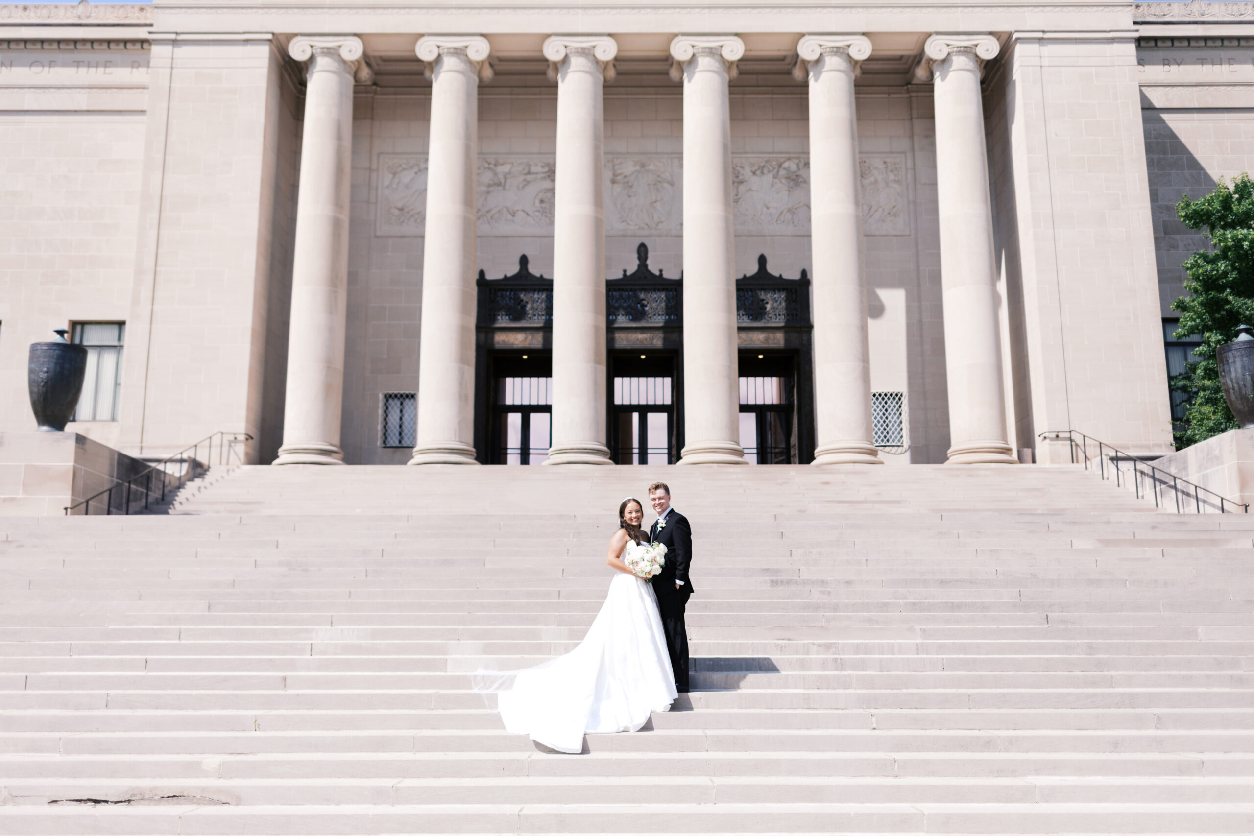Bride and groom standing on wide steps in front of a building with six tall columns.