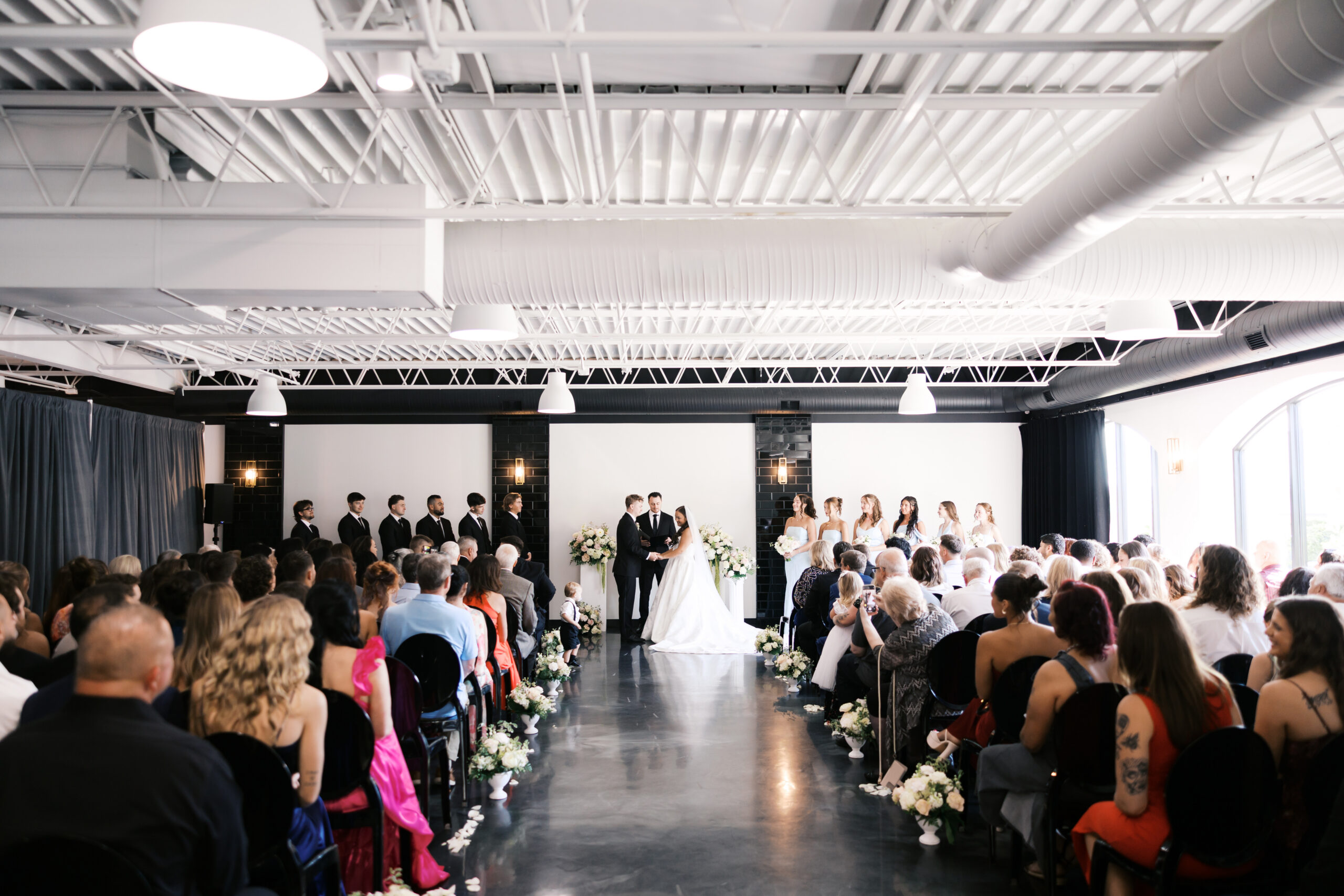 Wide shot of indoor wedding ceremony with guests seated in rows watching couple at altar.