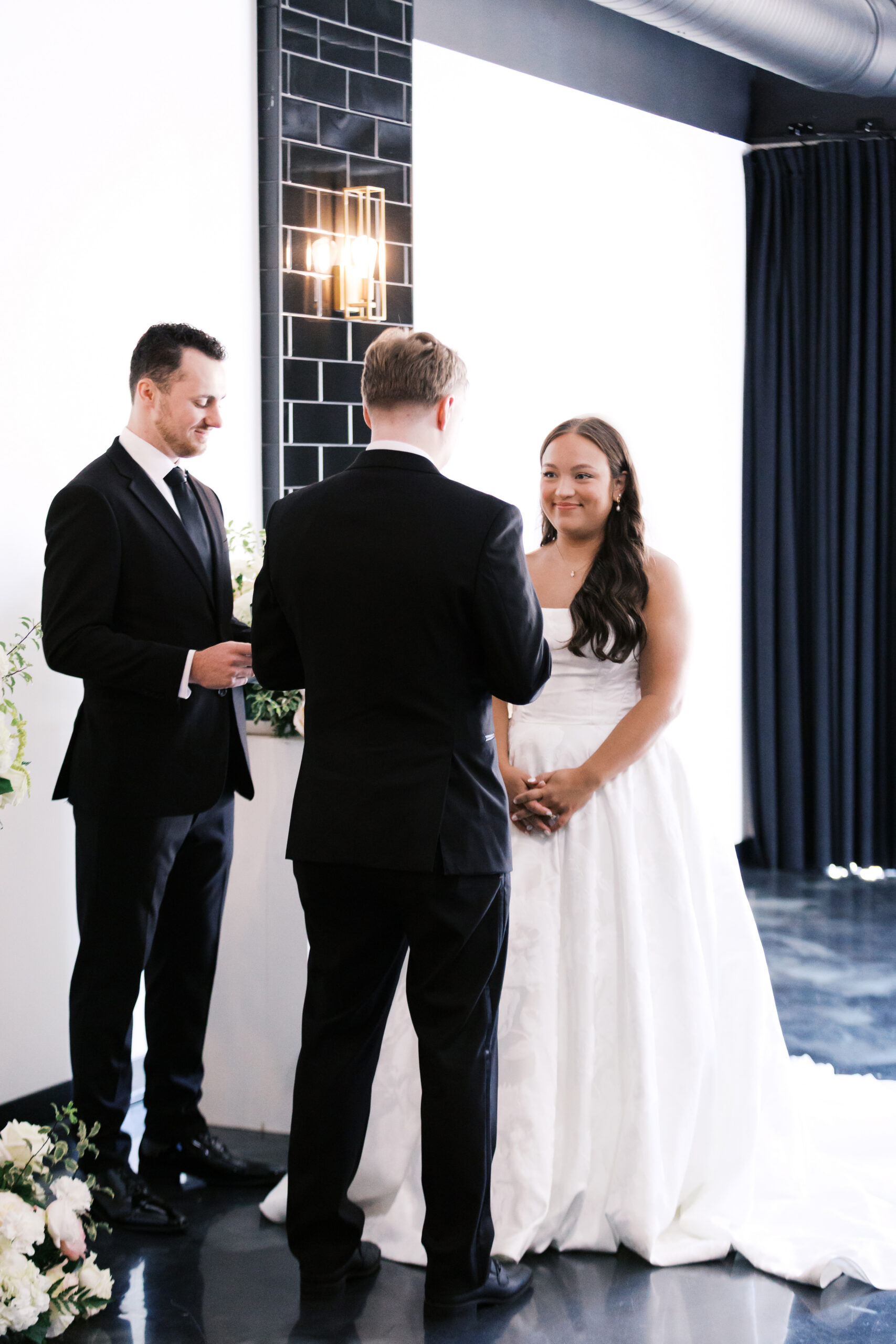 Bride smiling at groom during ceremony with officiant standing beside them.
