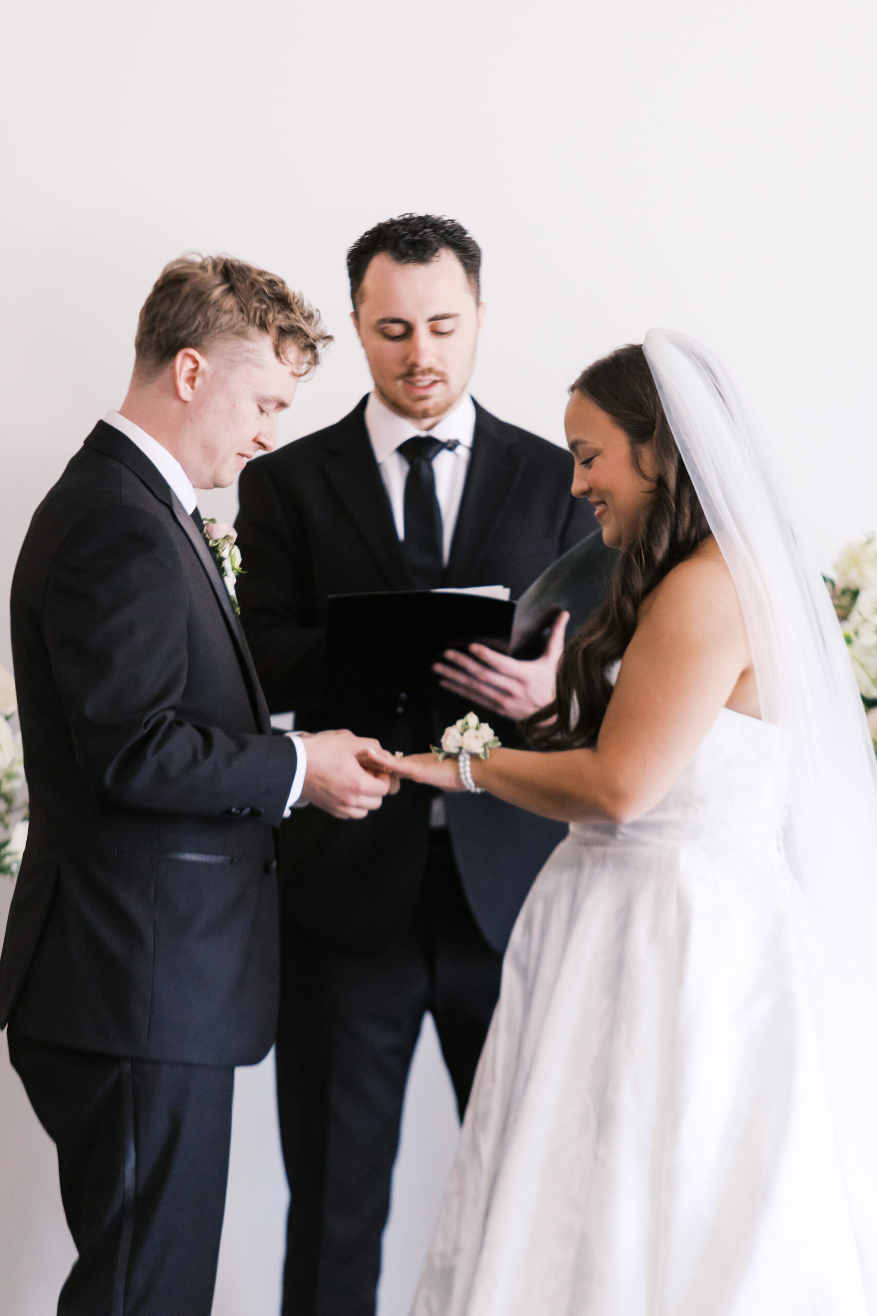 Groom placing ring on bride's finger during ceremony with officiant watching.