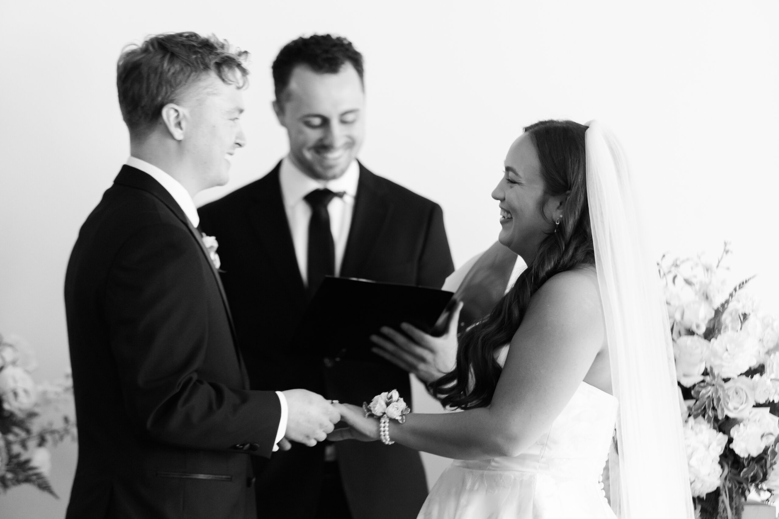 Black and white photo of bride and groom holding hands and smiling during ceremony.