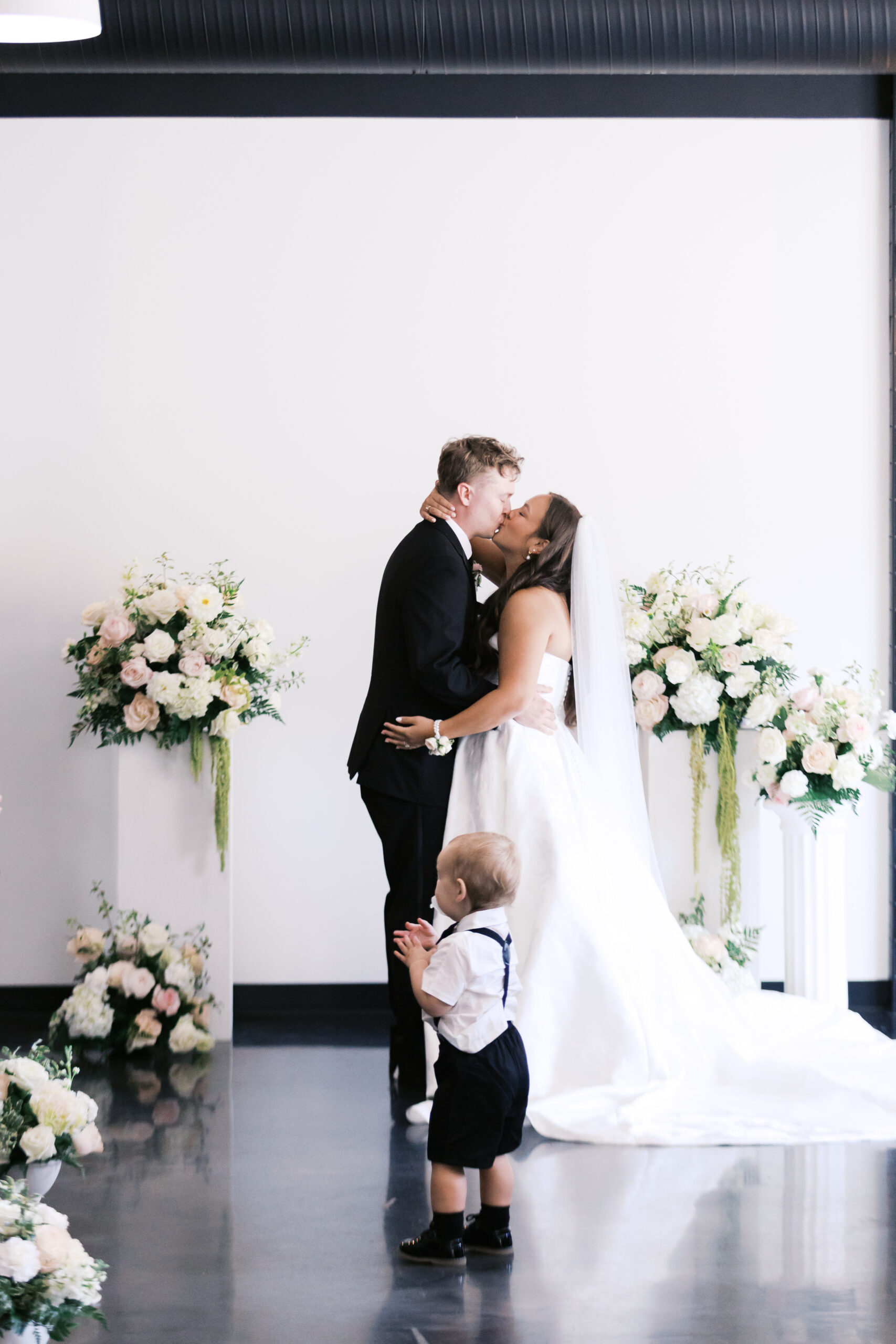 Bride and groom kissing at altar with toddler in formal wear standing nearby.