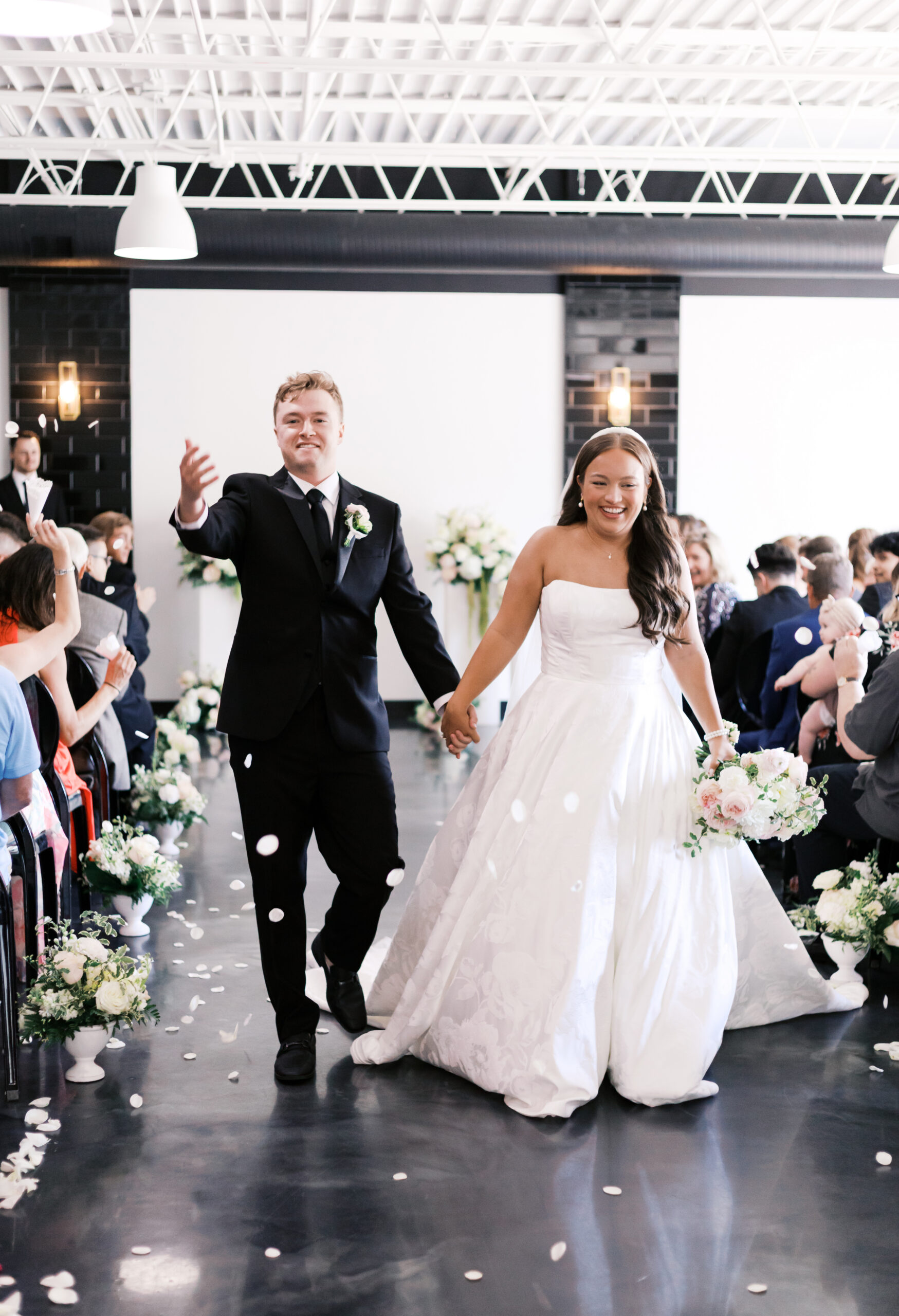Newlyweds walking down aisle holding hands with guests applauding and flower petals on floor.