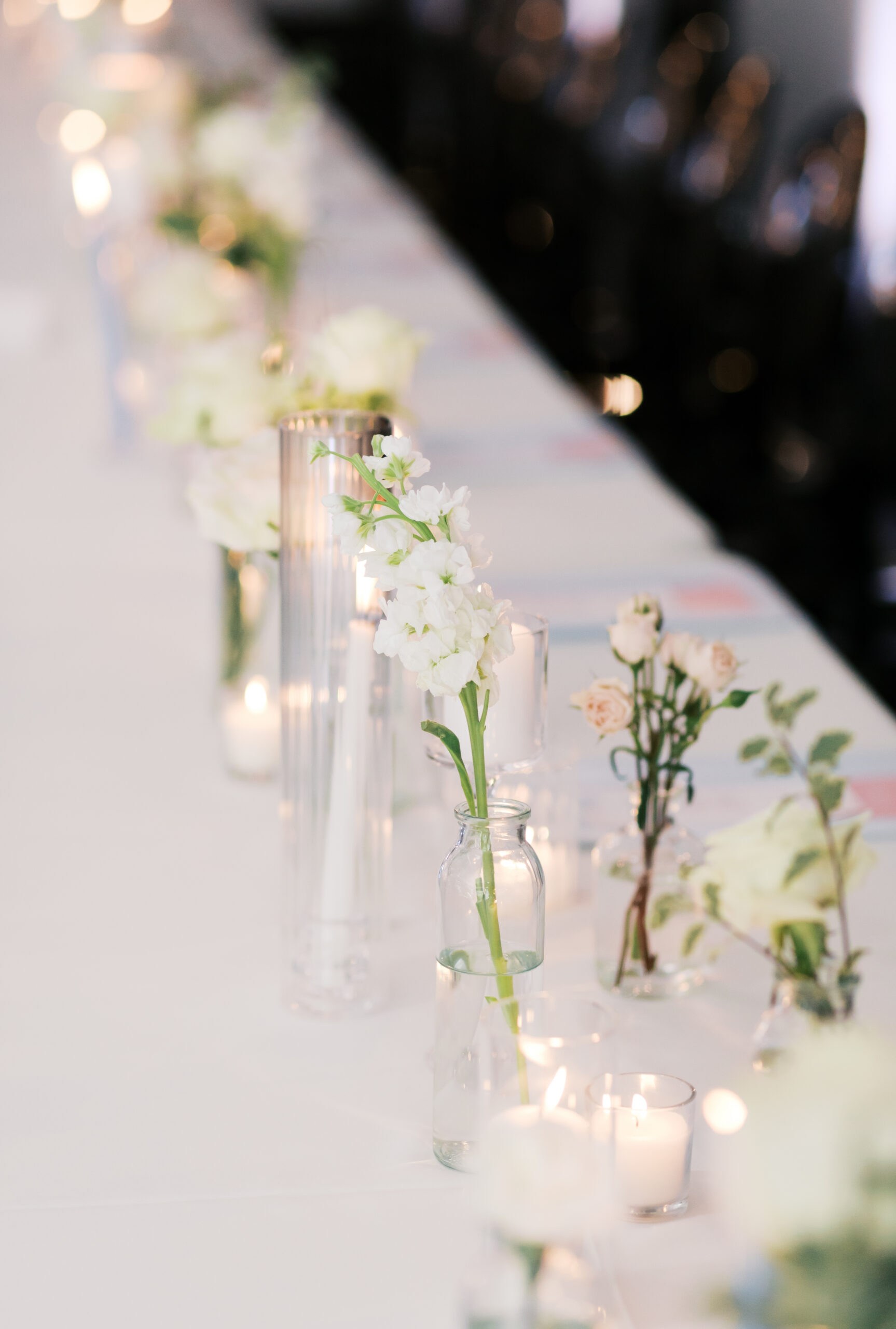 Row of small bud vases with white flowers and candles on long table.