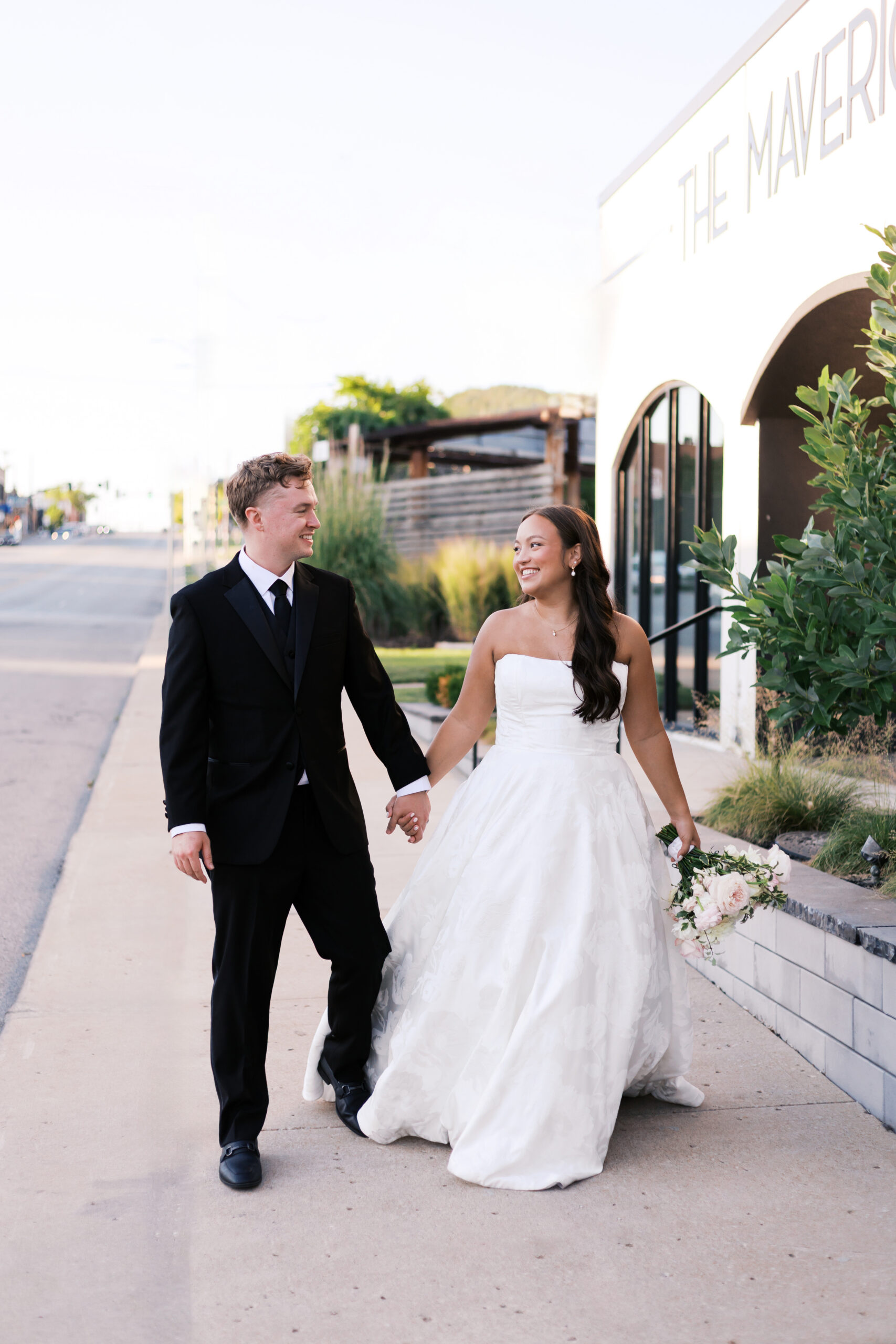 Bride and groom holding hands and looking at each other as they walk down a sidewalk in front of a building with large windows.