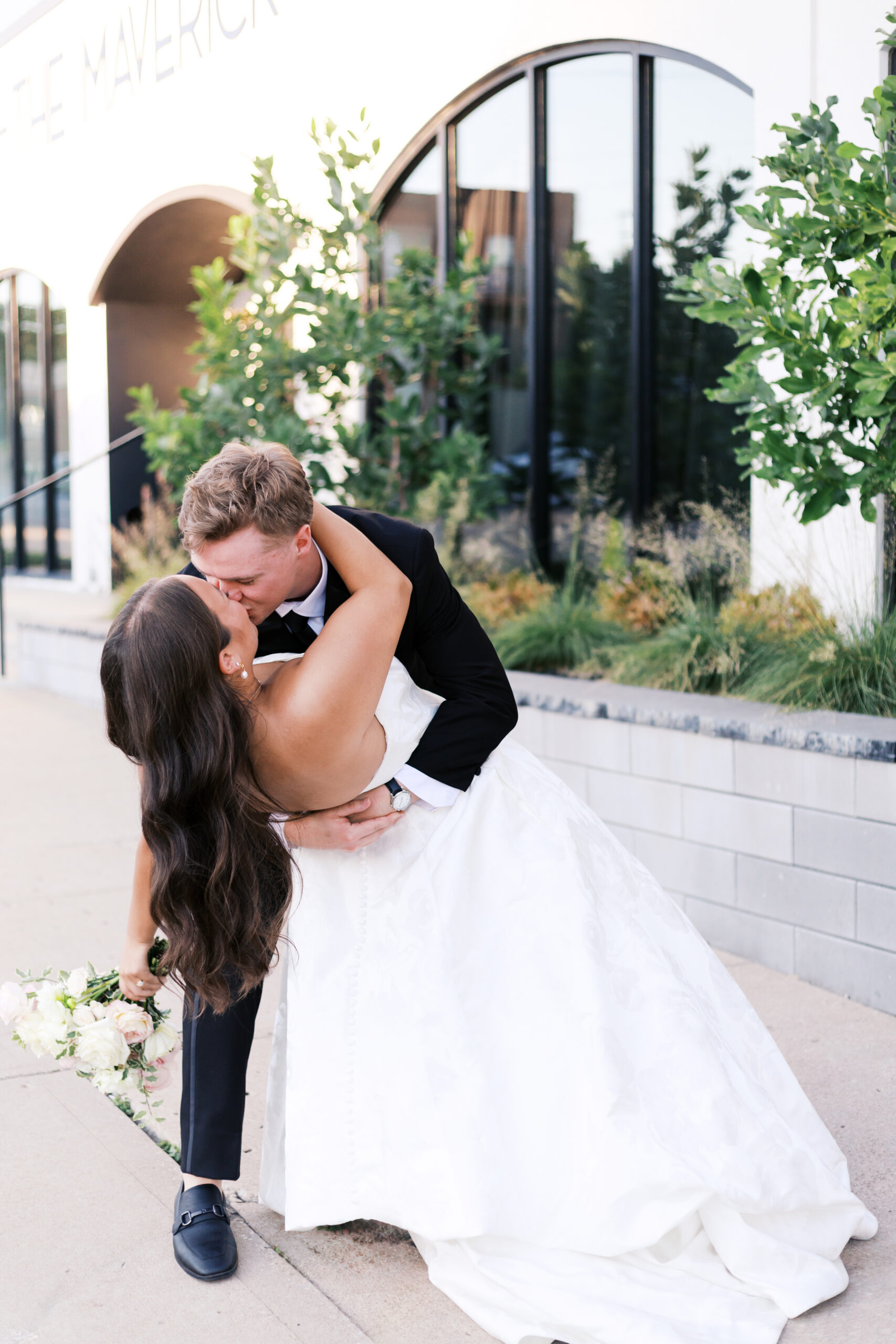Groom dipping bride as he kisses her on a sidewalk.