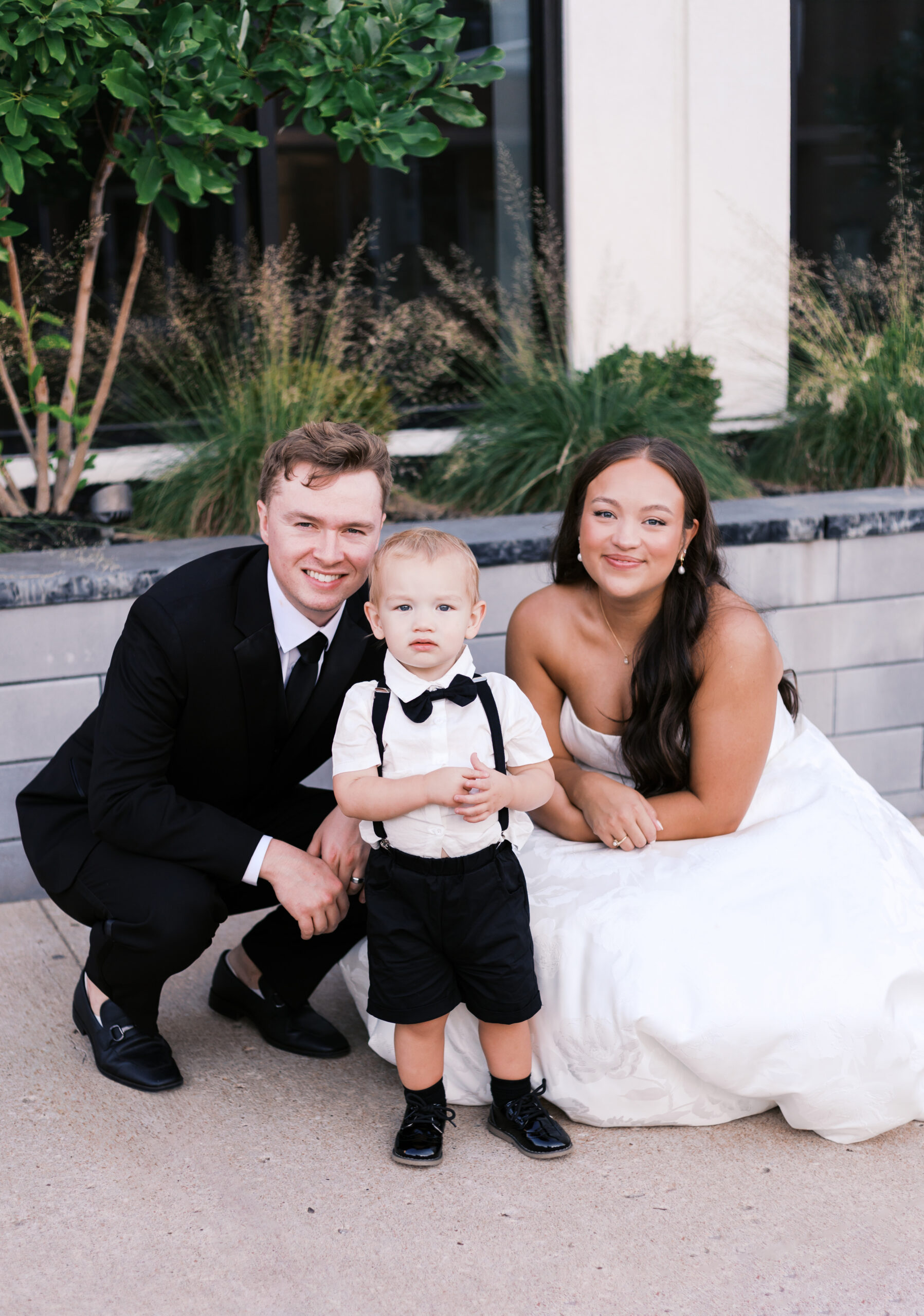 Bride and Groom kneeling with toddler in suspenders, bow tie, and shorts.