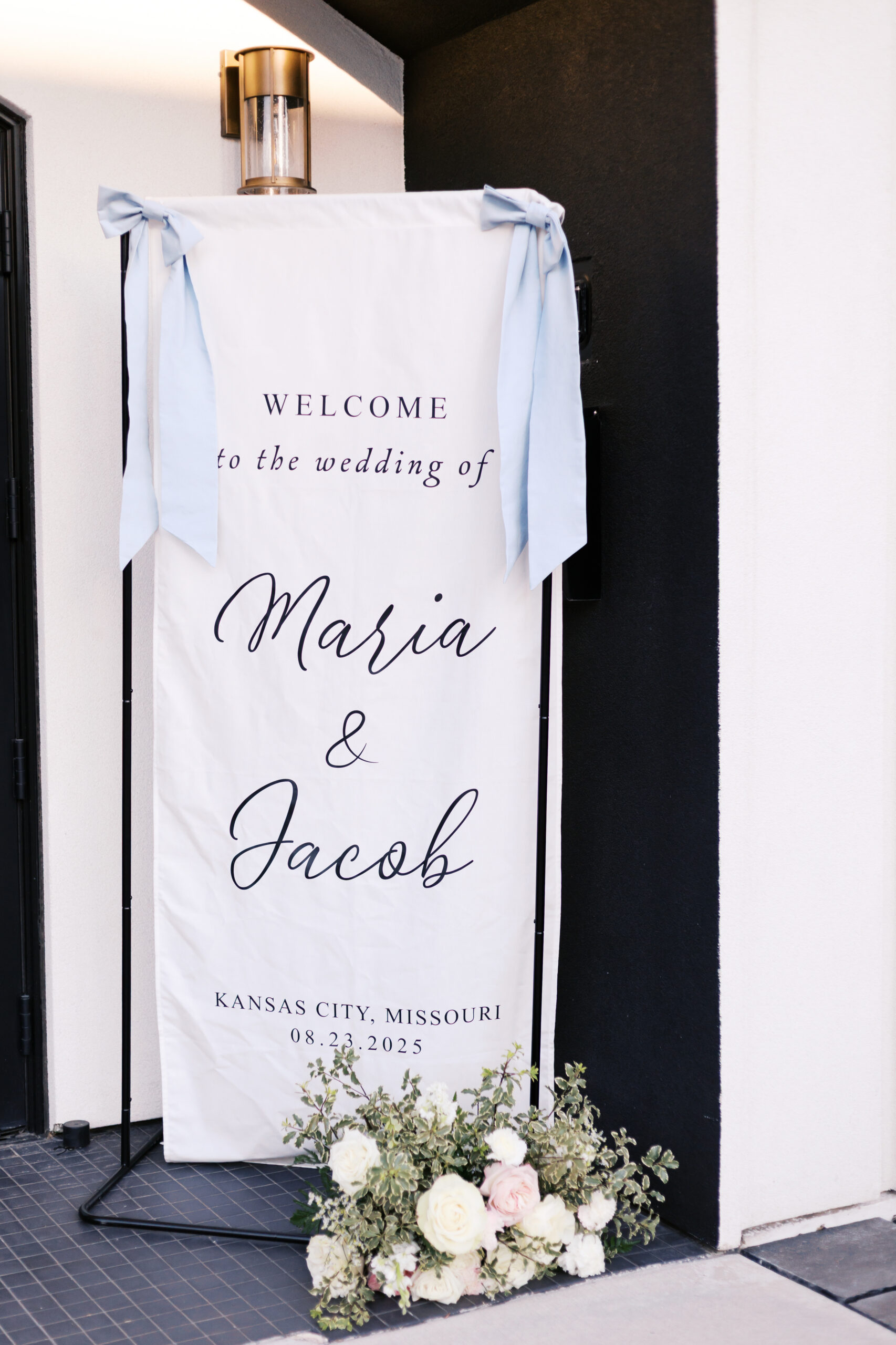 White fabric wedding welcome banner with couple's names hanging by entrance with light blue ribbon ties.