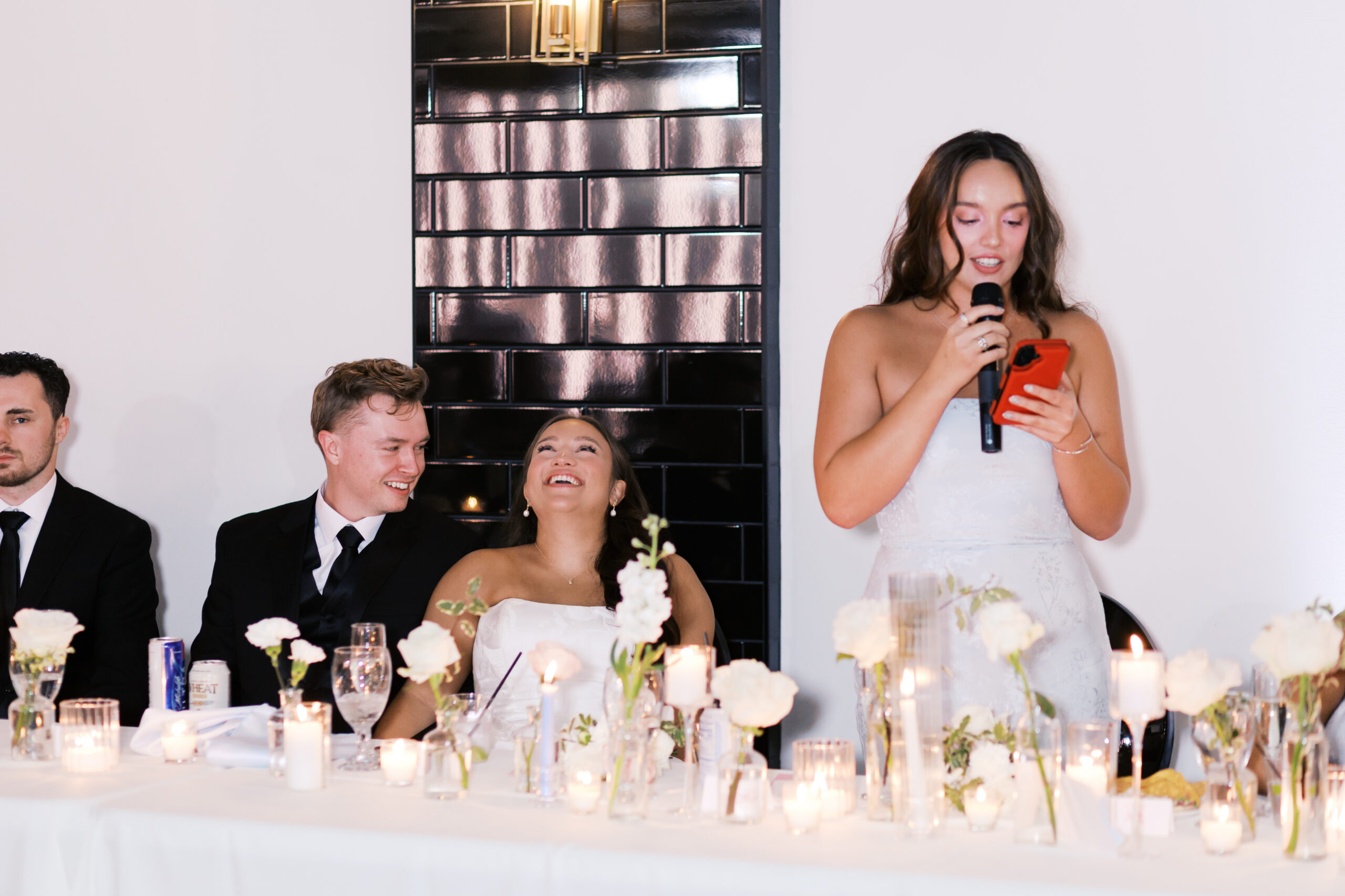 Bride and groom smiling and laughing as bridesmaid gives a speech.