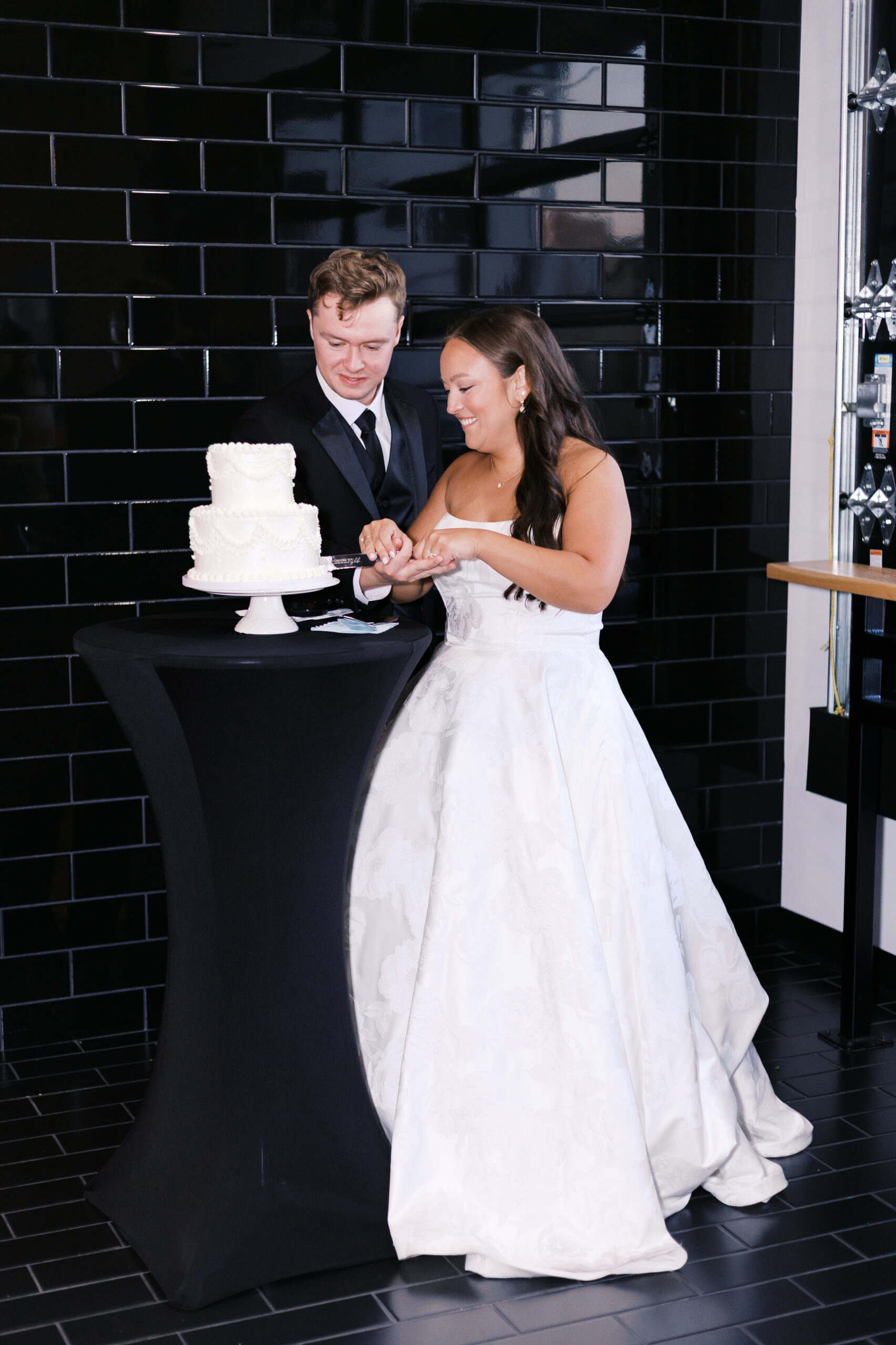 Bride and groom cutting a two tier wedding cake in front of a black wall.