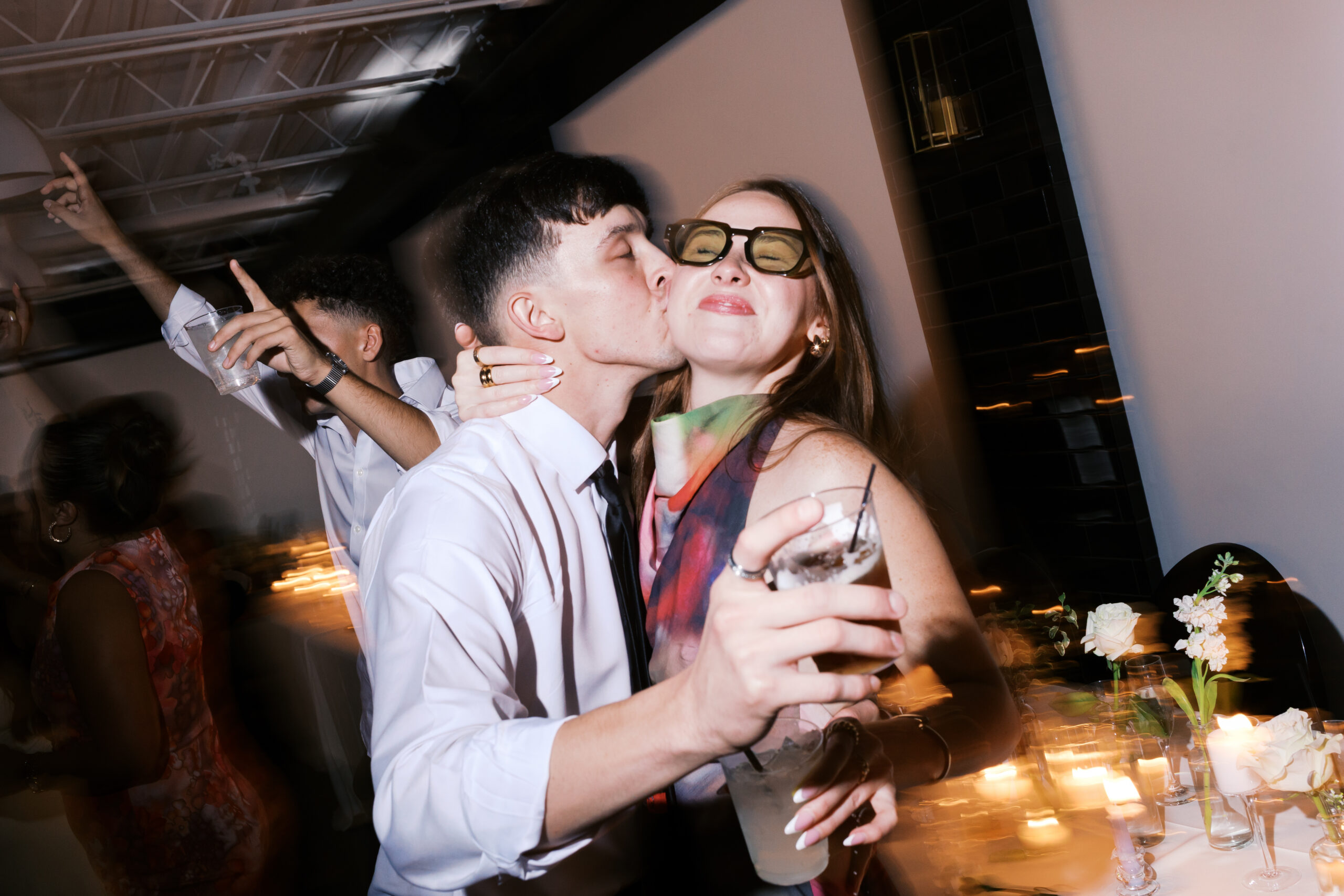 A man kissing a woman on the cheek at the reception as they are holding their drinks and the woman is smiling.