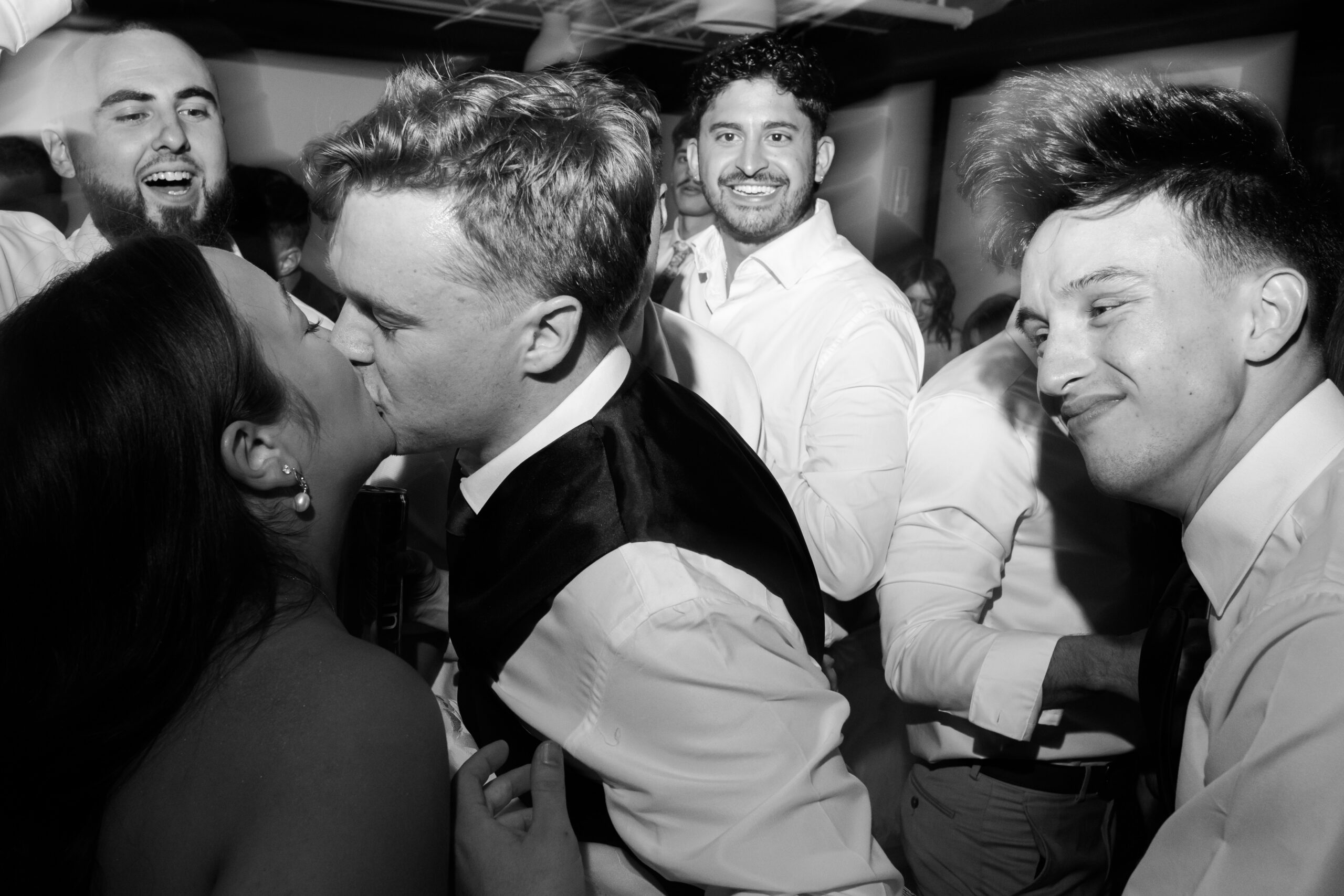Black and white photo of the bride and groom kissing in the foreground while people smile and dance behind them.