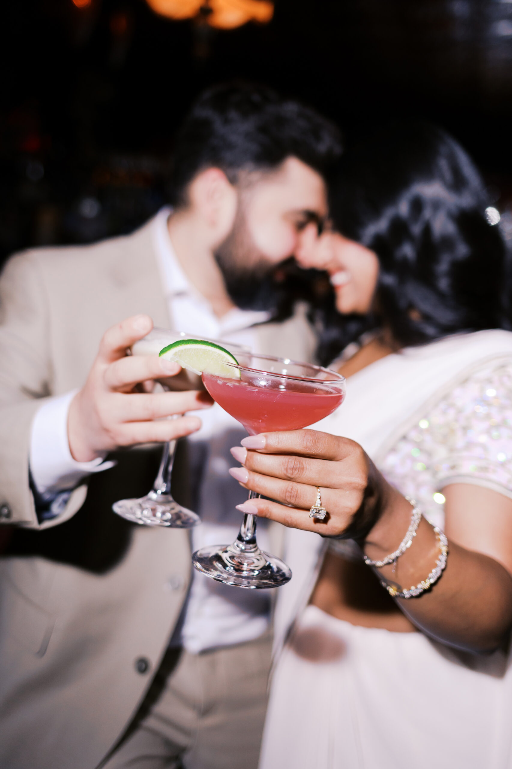 Close-up engagement photo of couple clinking cocktails with engagement ring visible, captured in a dark bar setting in Kansas City.