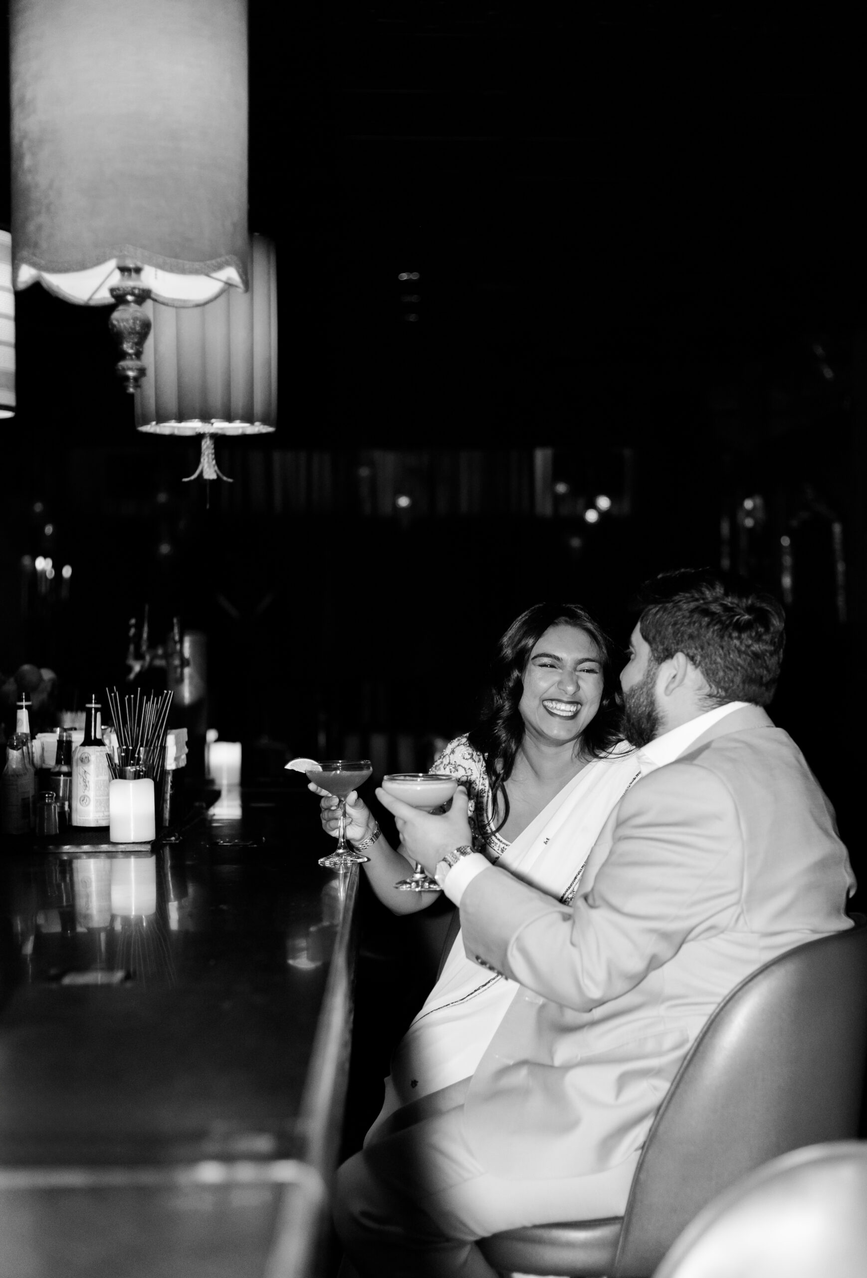 Black and white photo of couple laughing and toasting drinks at a dimly lit jazz bar engagement session in Kansas City.