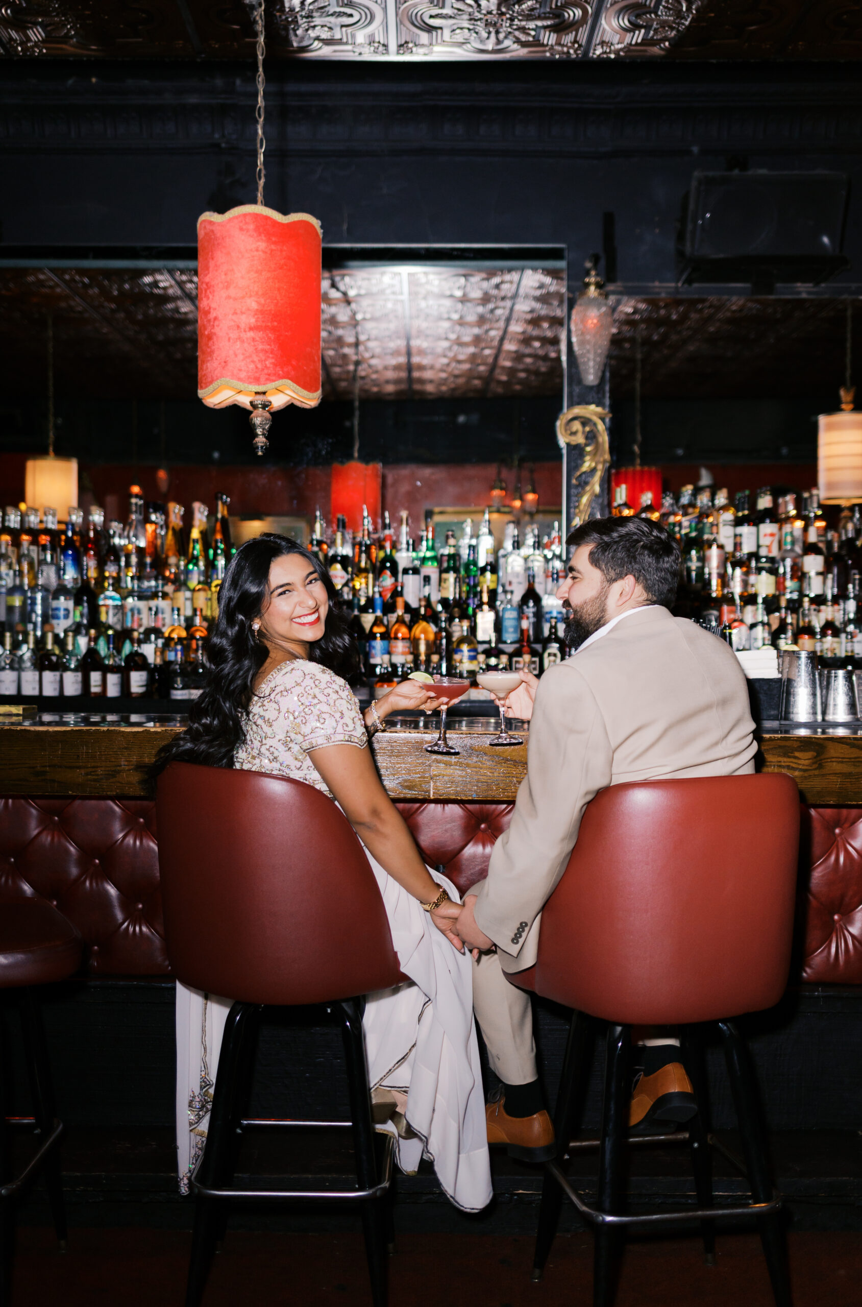 Engaged couple seated at the bar in Green Lady Lounge holding cocktails, surrounded by red walls and vintage lighting in Kansas City.