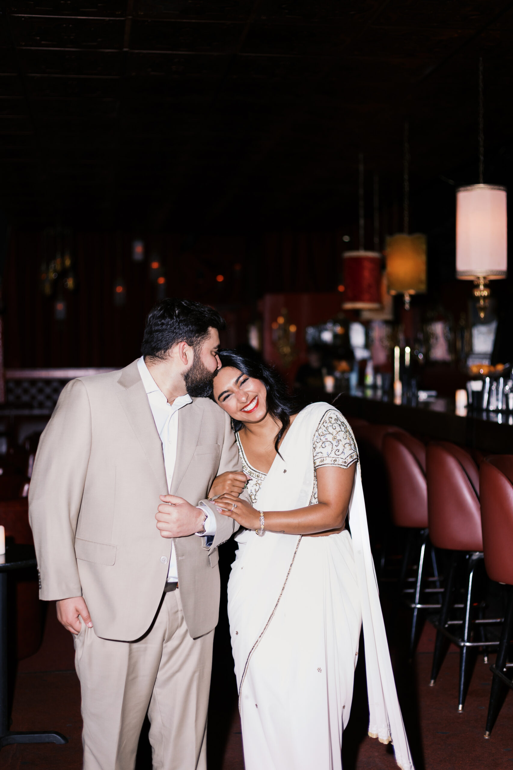 Man kissing his fiancée’s forehead inside Green Lady Lounge as she smiles, wearing a white saree during their Kansas City bar engagement photos.
