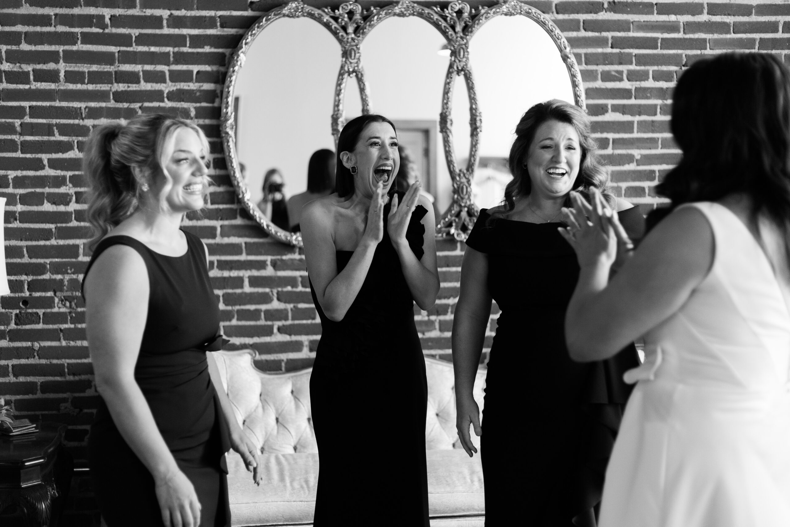Bridesmaids reacting with surprise and smiles as they see the bride in her dress in a brick loft room.