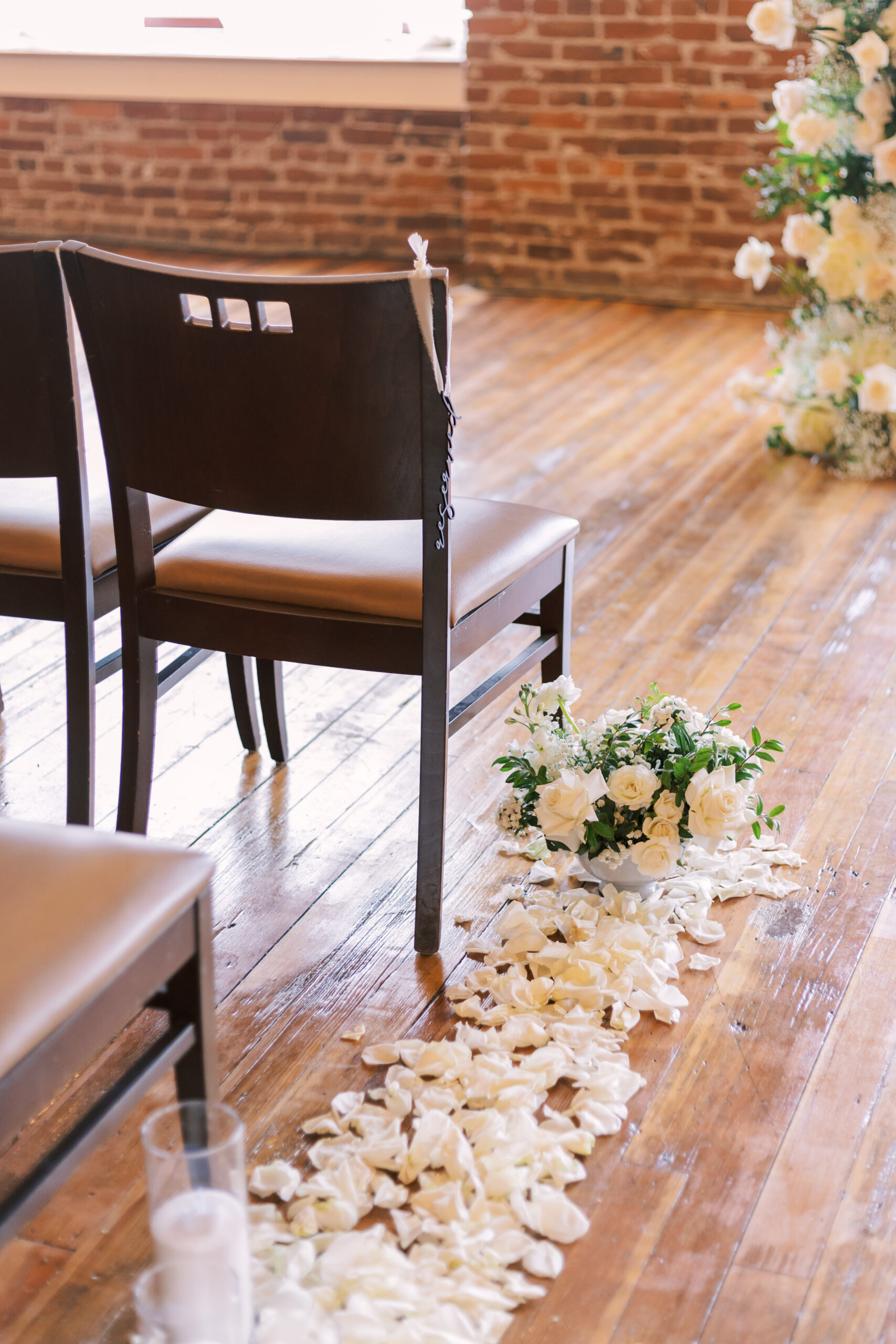 Wooden ceremony chairs beside an aisle lined with white flower petals and floral arrangements.