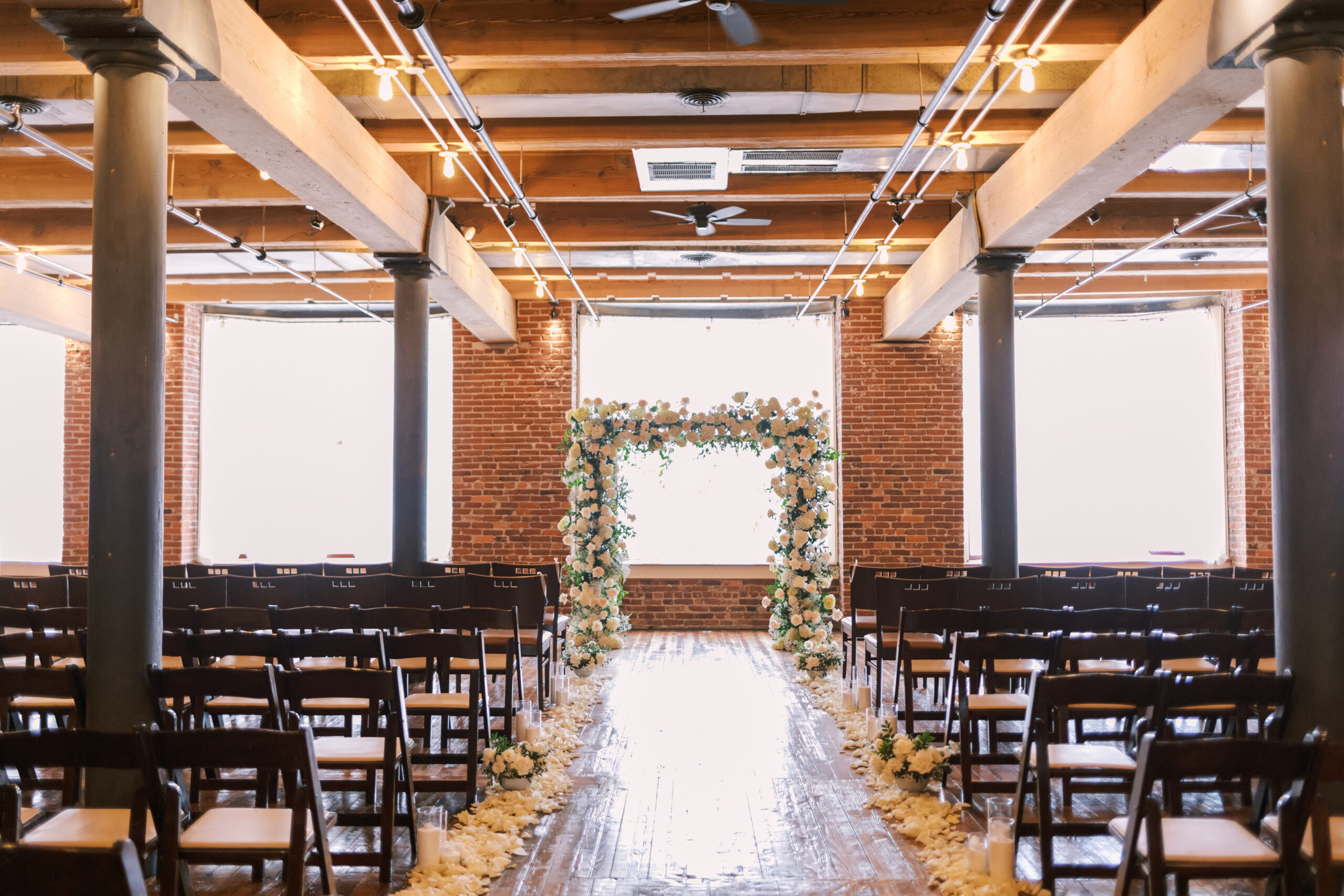 Wedding ceremony setup with rows of wooden chairs and a floral arch in a bright brick loft venue.