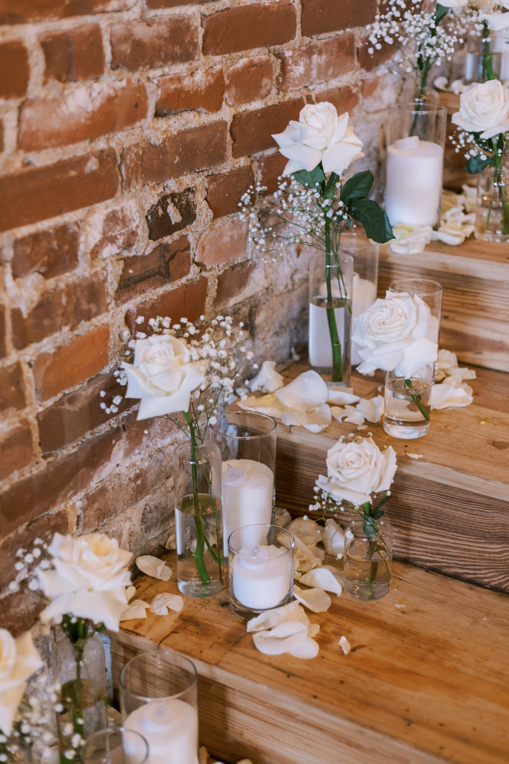 White roses and candles arranged in glass vases along wooden steps against a brick wall.