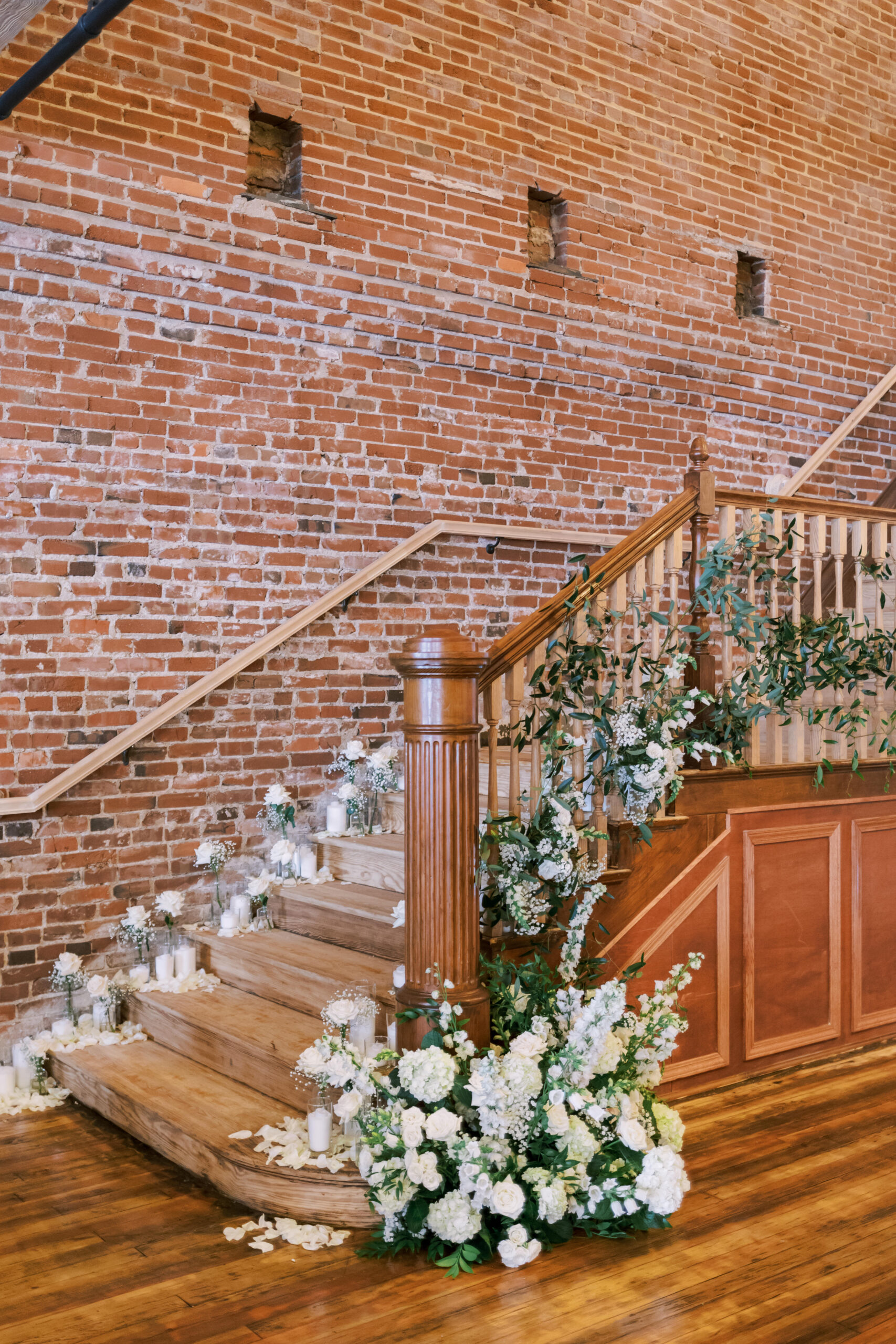 Wood staircase decorated with white florals, greenery, candles, and scattered petals in a brick venue.