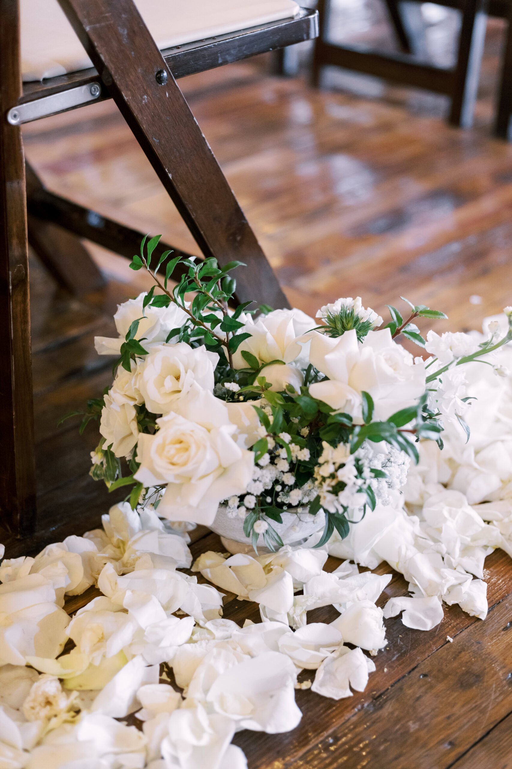 Low arrangement of white roses and greenery placed beside a ceremony chair with scattered petals.