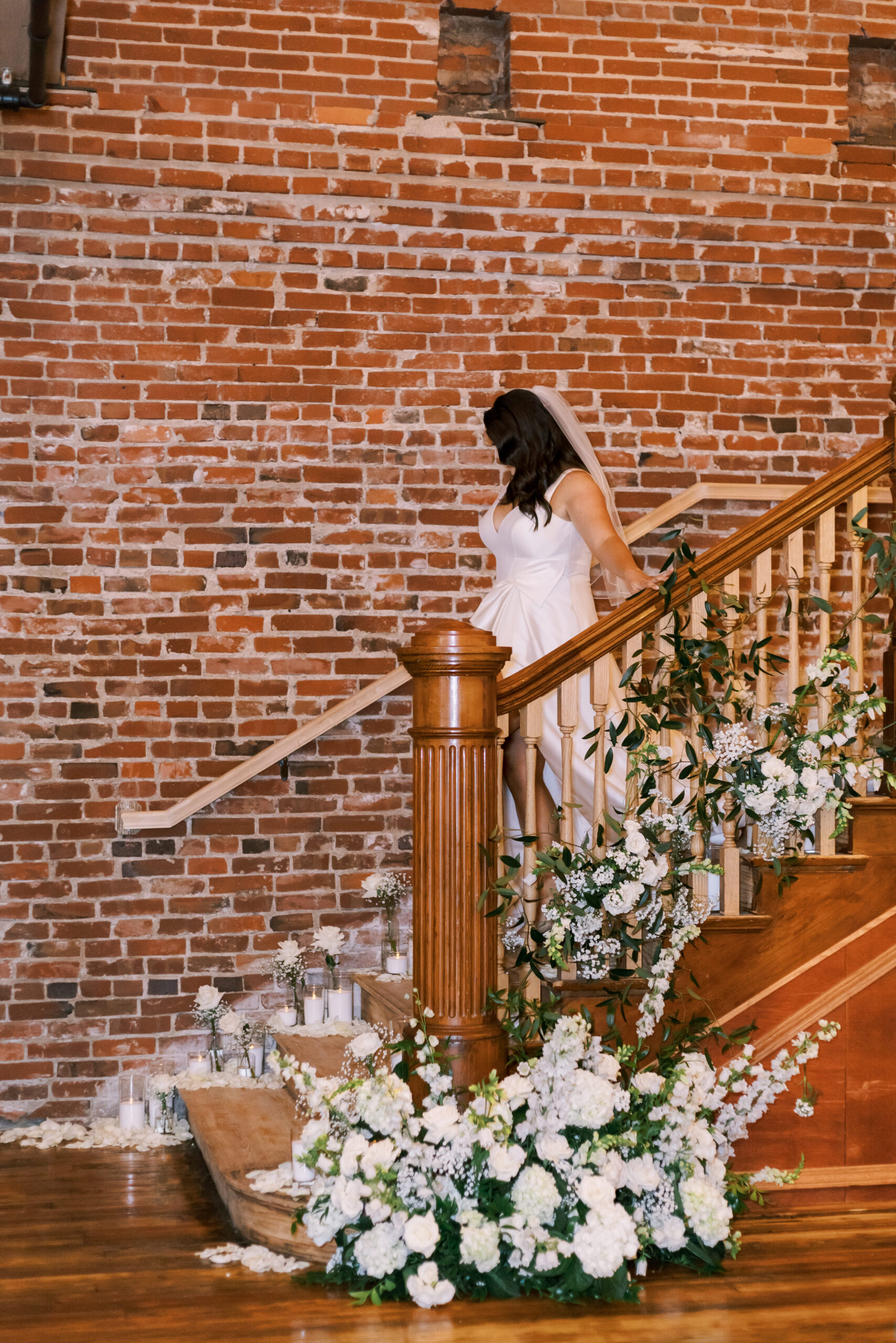 Bride in a white gown descending a wooden staircase decorated with white flowers and greenery.