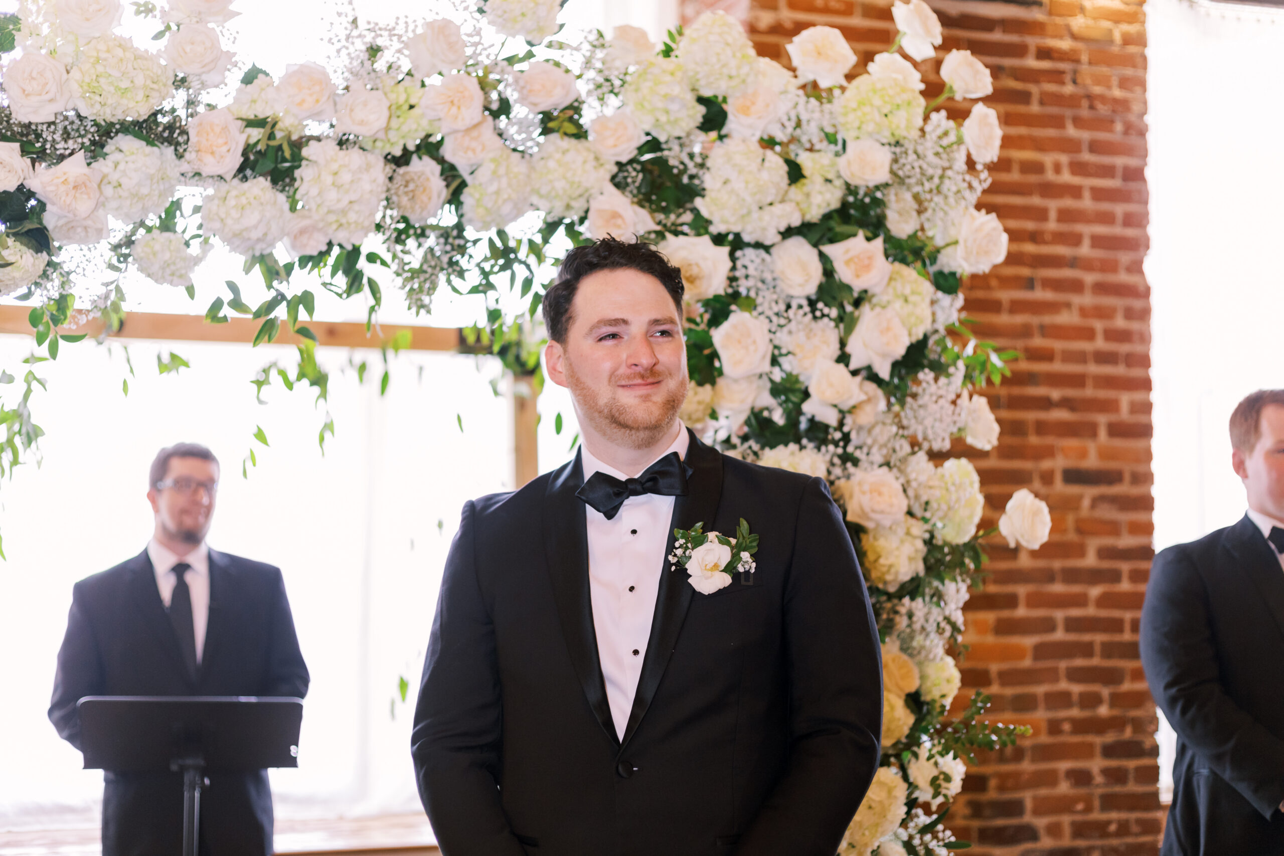 Groom in a black tuxedo standing beneath a white floral ceremony arch as guests arrive.