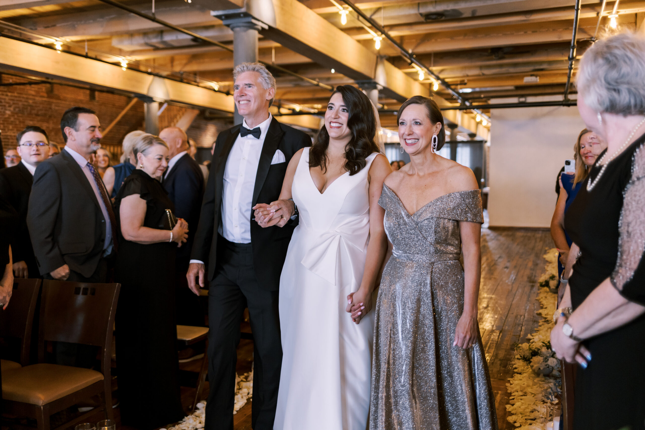 Bride walking down the aisle between two family members during the ceremony in a brick loft venue.