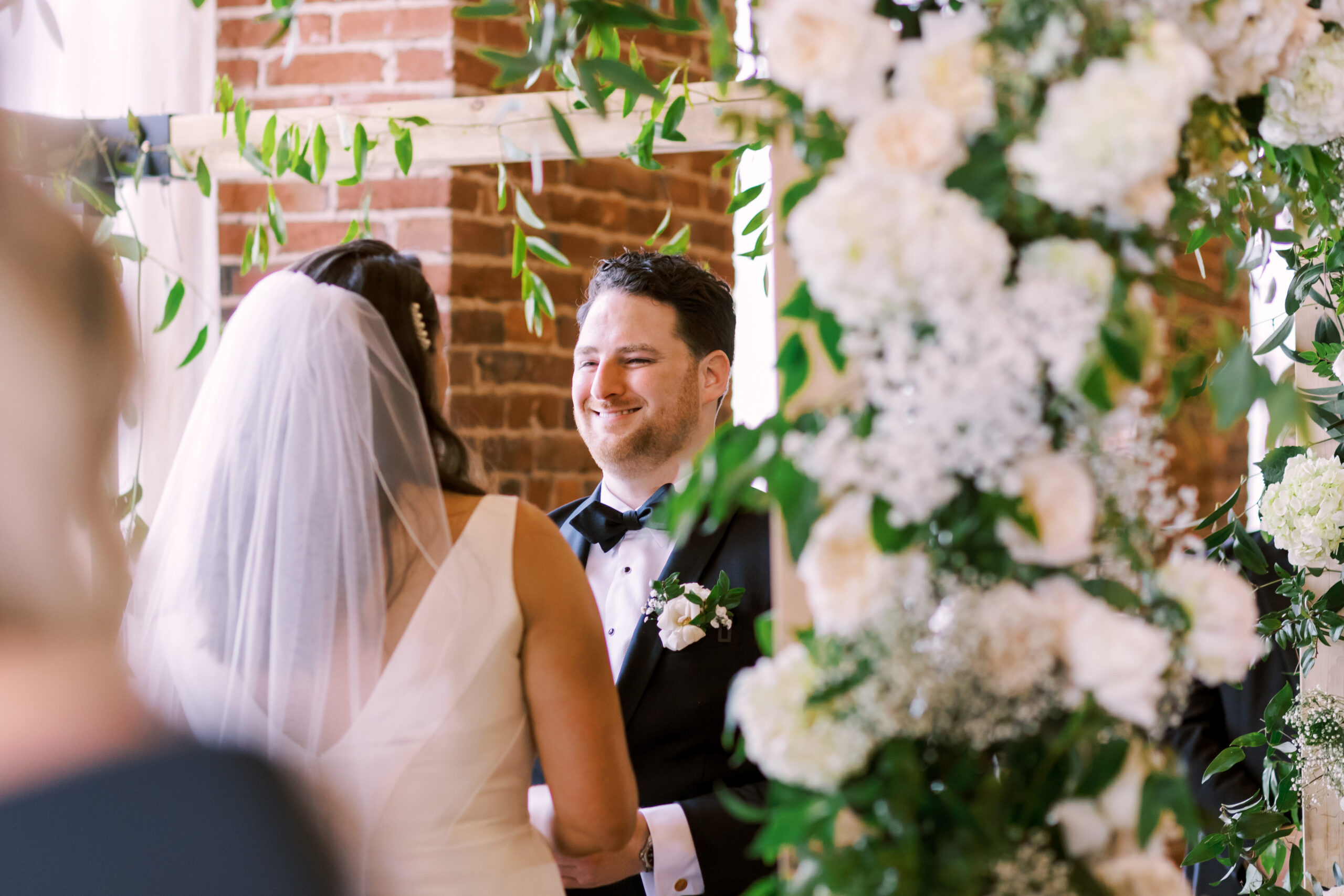 Groom smiling at the bride as they stand together during their ceremony beneath a floral arch.