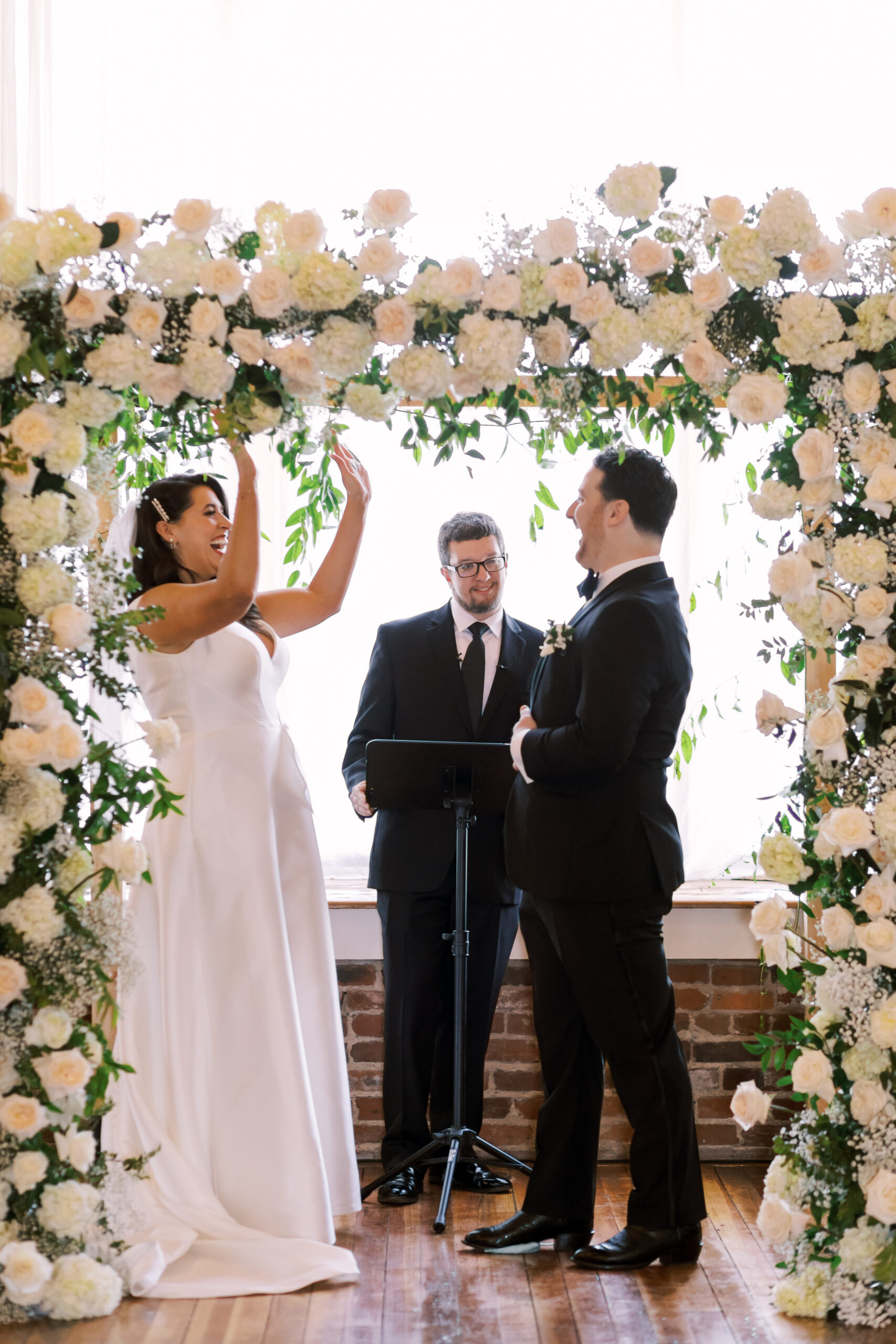 Bride raising her hands and laughing with the groom after exchanging vows under the floral arch.
