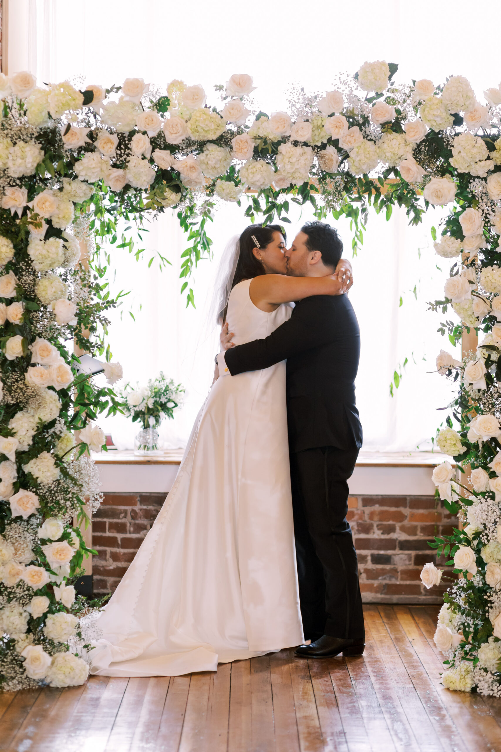 Bride and groom kissing beneath a large white floral ceremony arch.