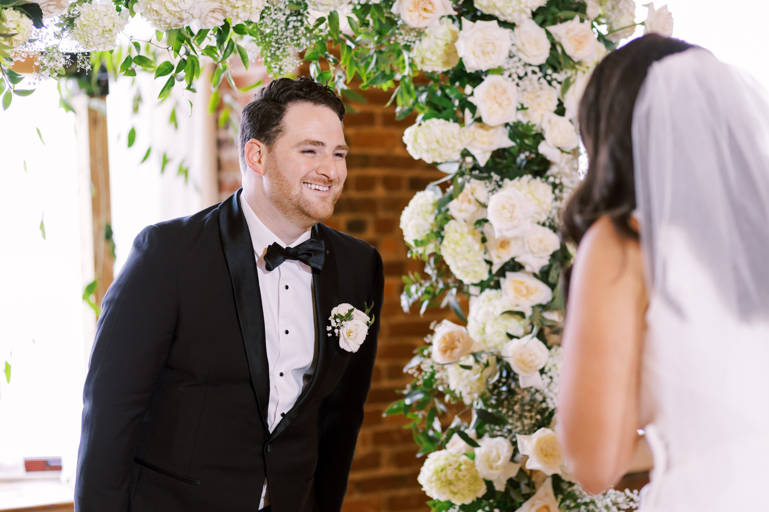 Groom smiling at the bride during the ceremony beneath a white floral arch.