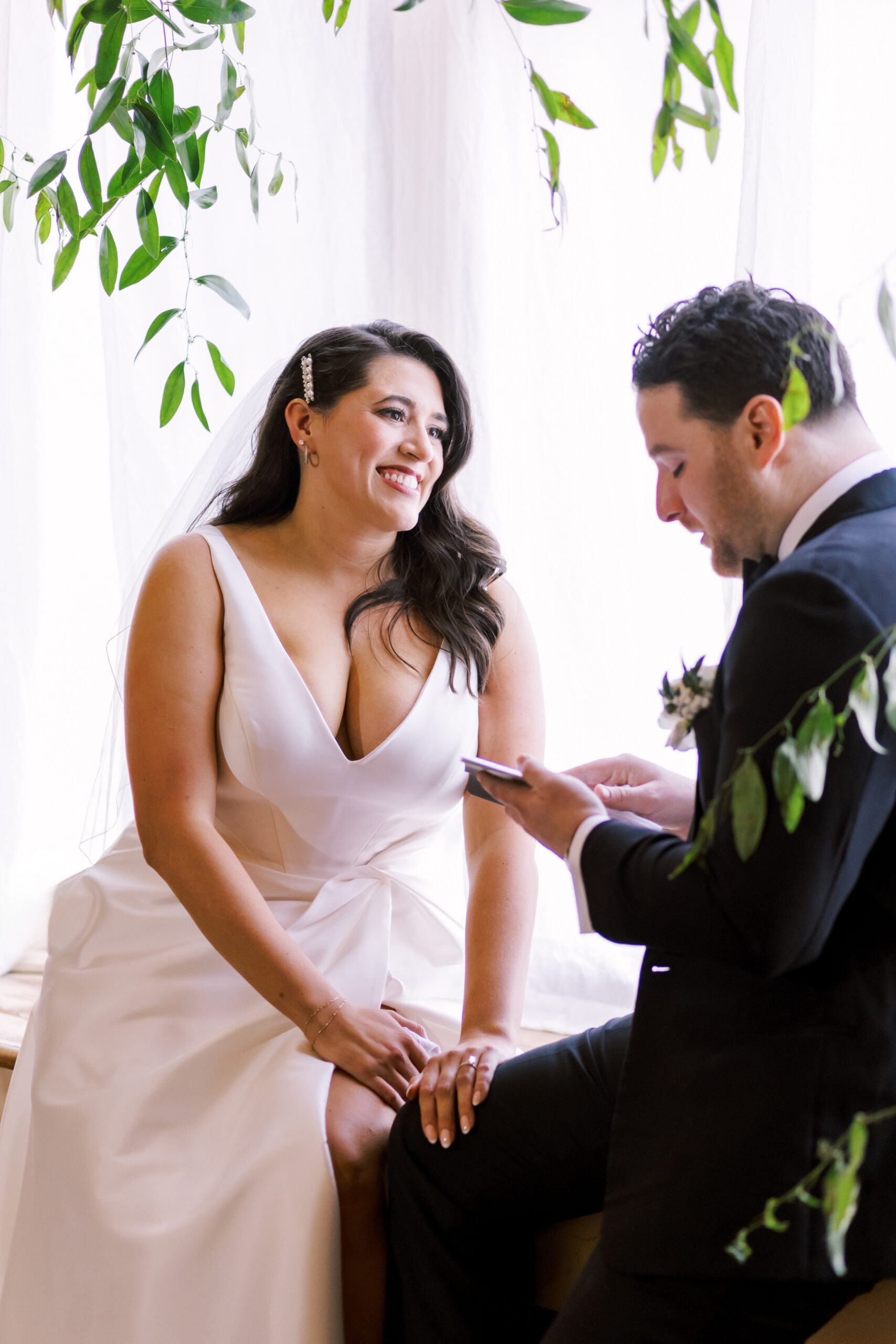 Bride seated and smiling while the groom reads his vows beneath greenery and soft window light.