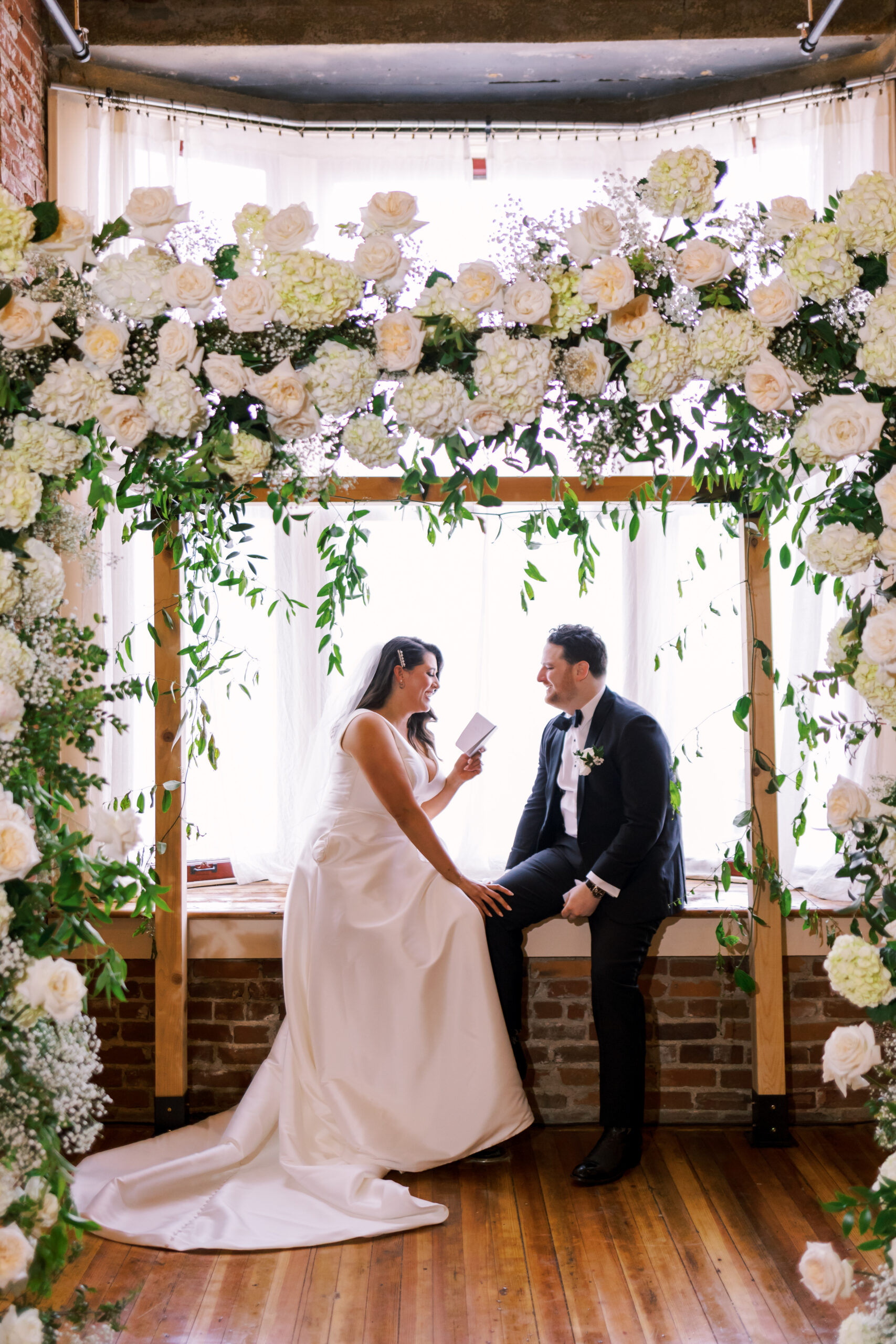 Bride and groom seated beneath a white floral arch reading handwritten vows to each other.