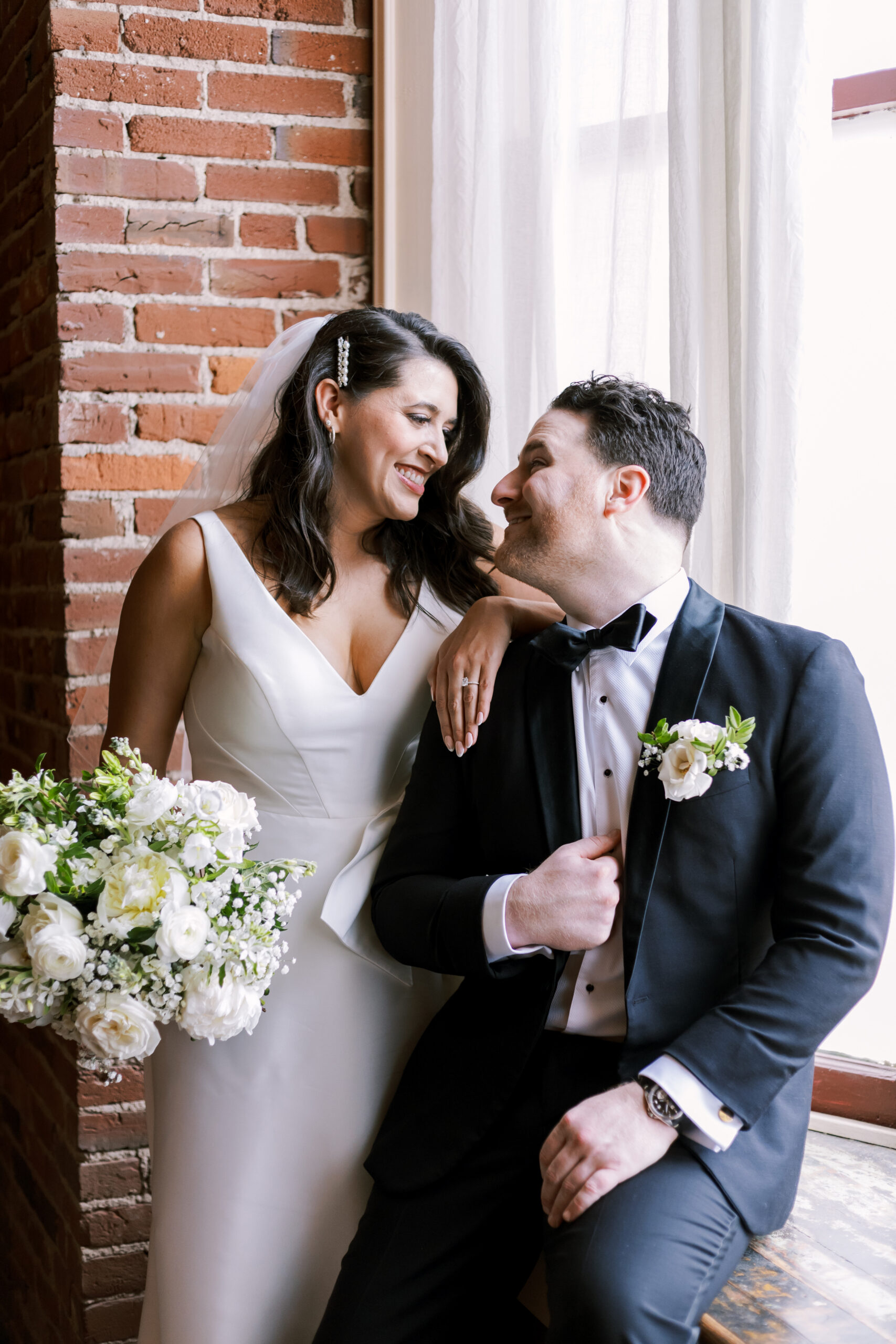 Bride holding a white bouquet while leaning beside the groom near a large window and brick wall.