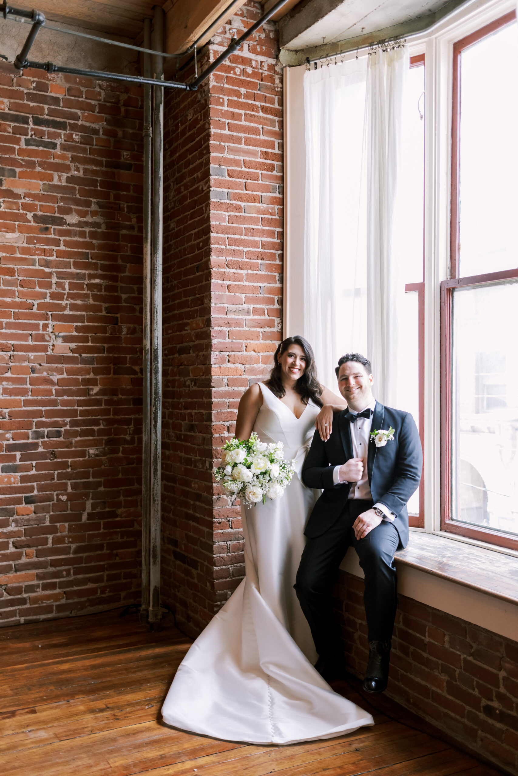 Bride and groom posing together beside a tall window in a brick loft wedding venue.